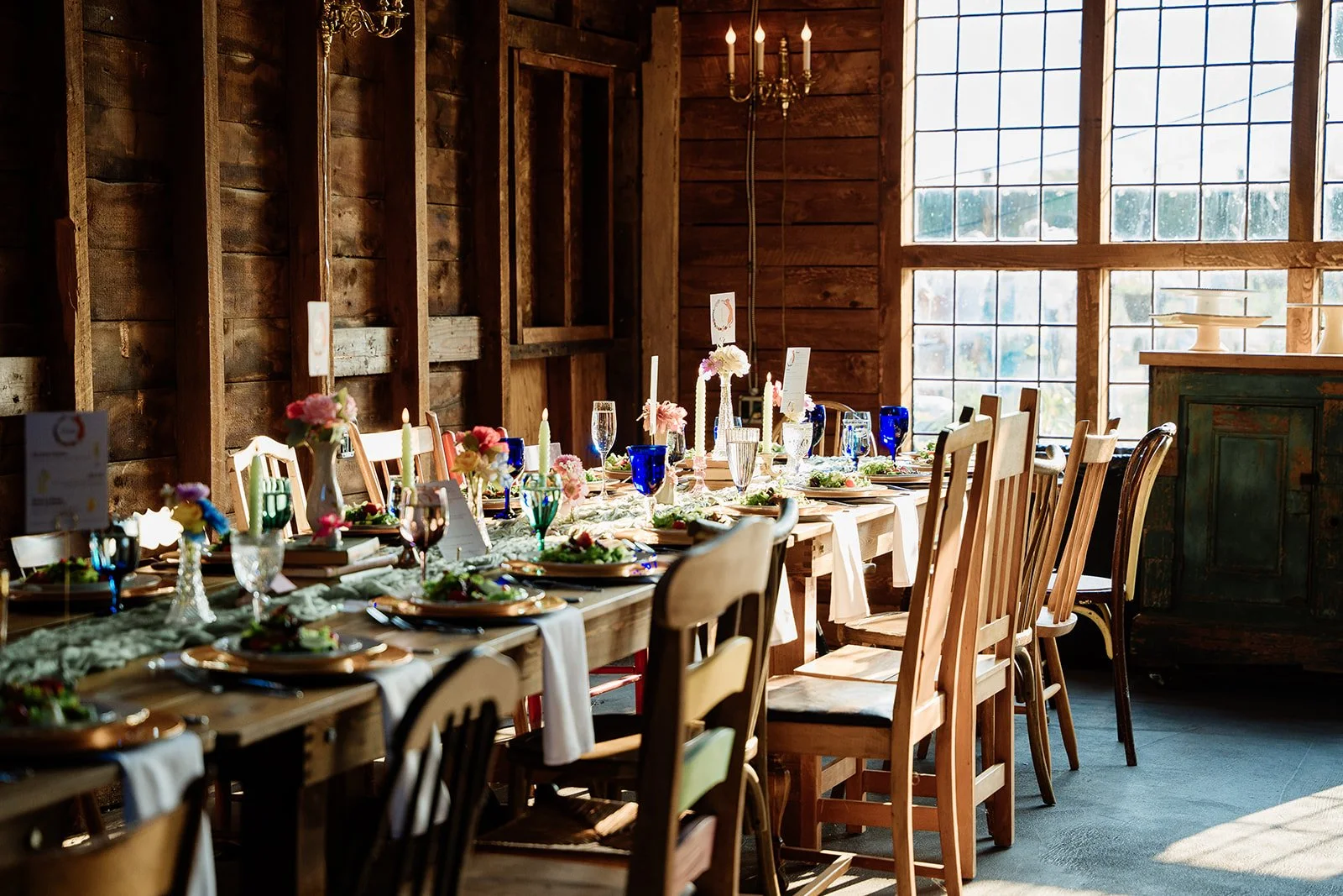 A rustic indoor dining table set for a celebration with floral centerpieces, candles, and colorful glassware, in a warmly lit wooden room with large windows.