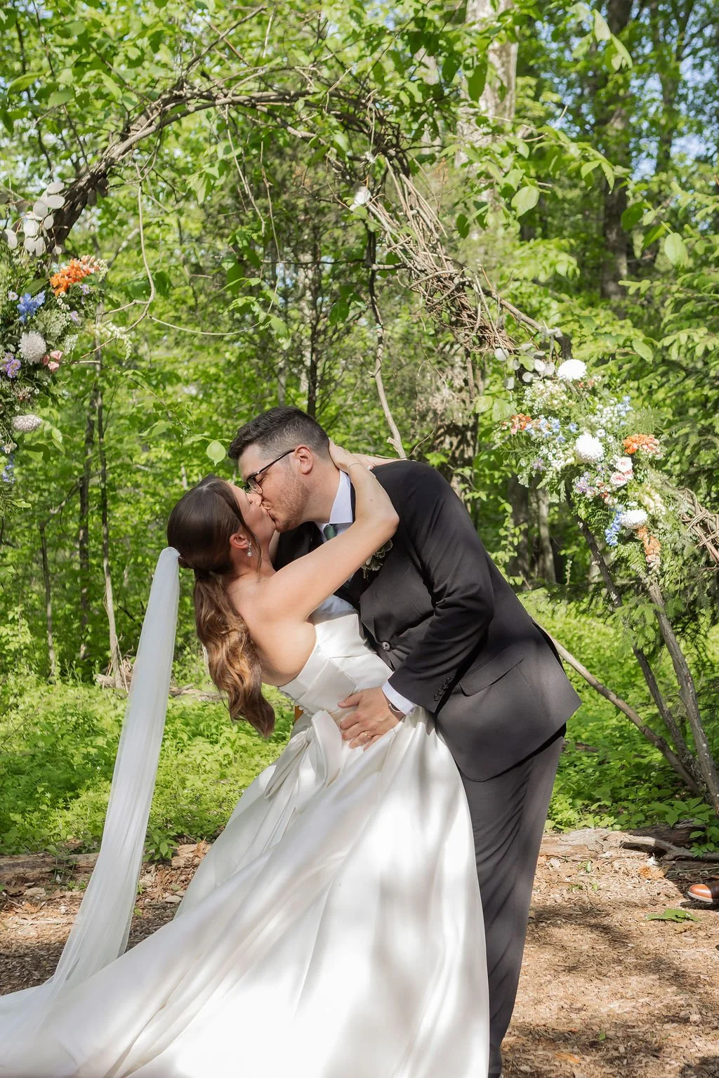 A newlywed couple sharing a kiss during their wedding ceremony outdoors in a green forest, under a natural arch decorated with flowers and vines.