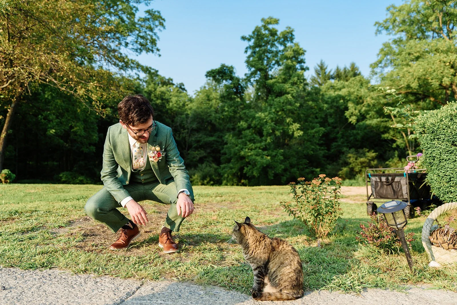 A groom in a green suit reaches out to pet a barn cat at Catskills wedding venue Black Walnut Farm