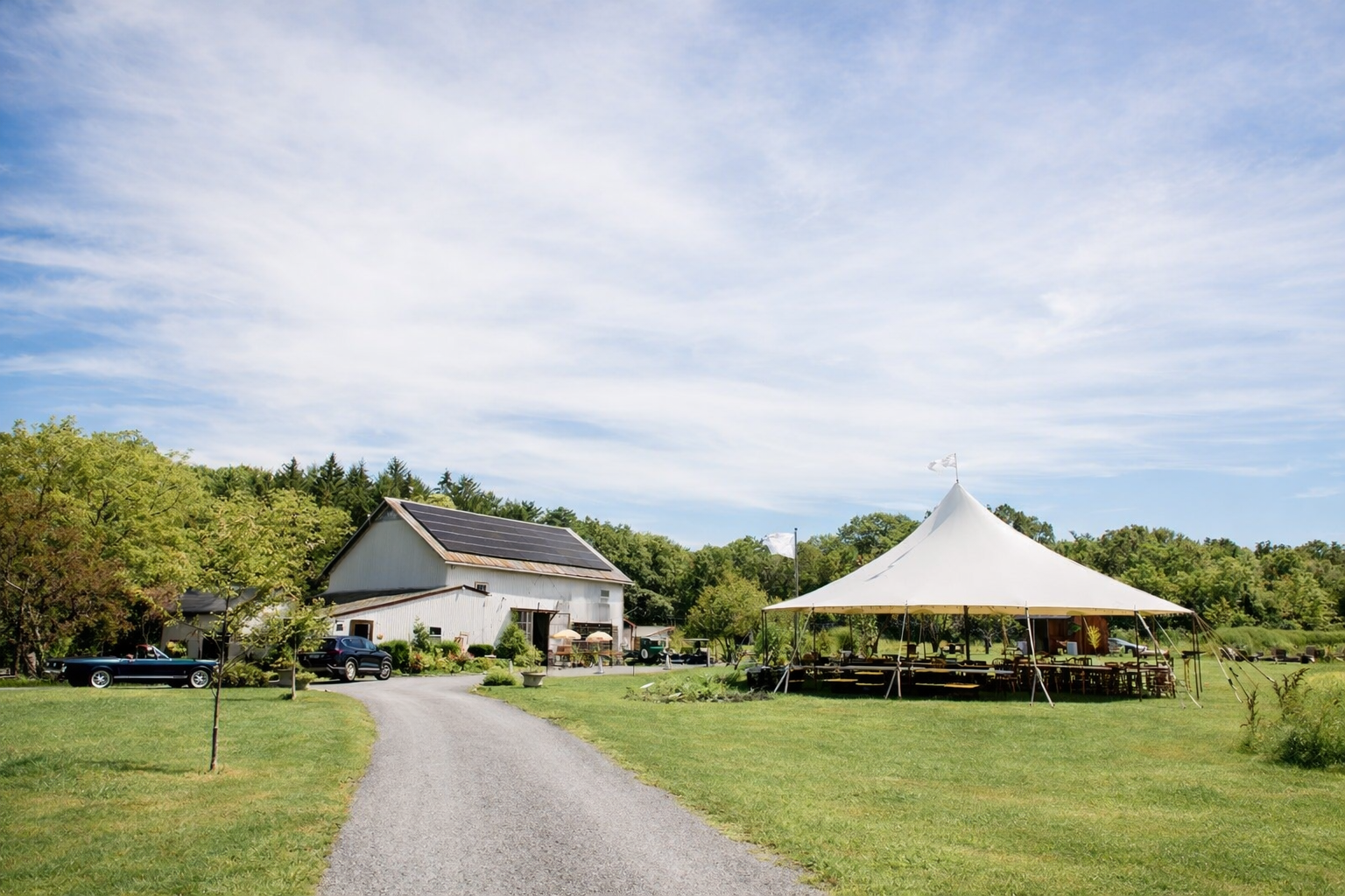 A rural outdoor scene with a gravel driveway leading to a white barn with solar panels on the roof. To the right, there is a large white tent set up on the green grass with picnic tables inside. Trees and a partly cloudy sky are in the background.