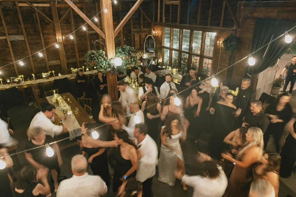 Guests dancing at a wedding reception in a rustic barn, with string lights overhead, tables decorated with flowers, and a large window allowing natural light in.
