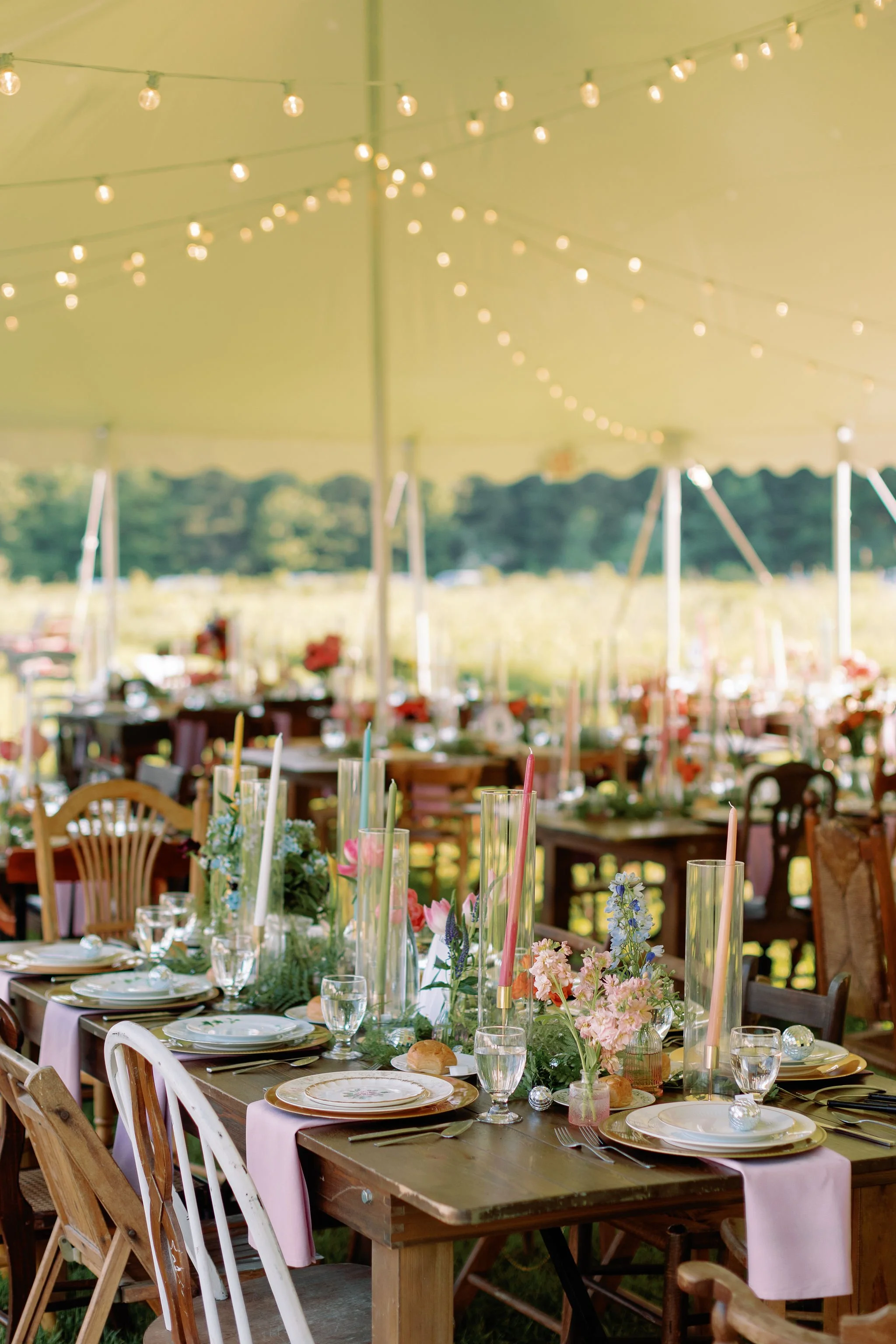 Decorated outdoor wedding reception under a beige tent with string lights, featuring a wooden table with pastel pink table runner, tall candles, floral arrangements, dinner plates, glasses, and silver ornaments.