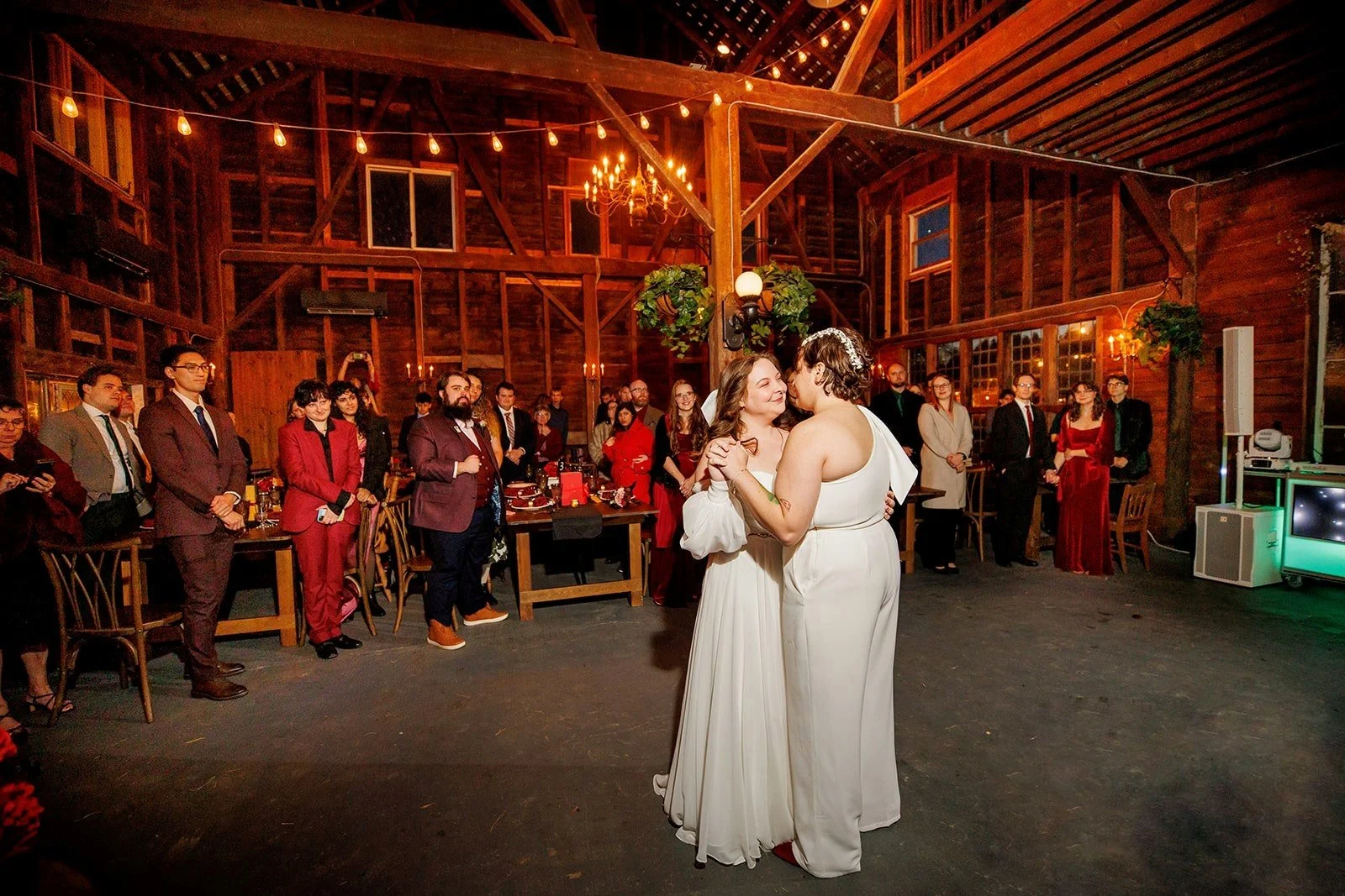 Two women in white dresses share a dance in a rustic barn decorated with warm lighting and string lights during a celebration, with guests watching in the background.
