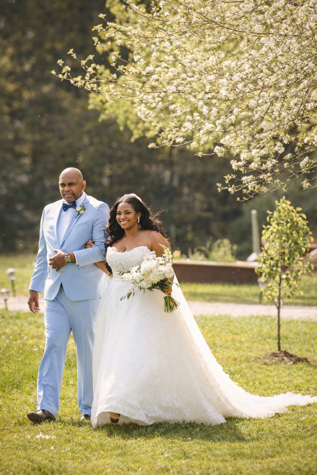 A bride in a white wedding gown holding a bouquet walking with a man in a light blue suit outdoors on a sunny day, surrounded by trees and greenery.