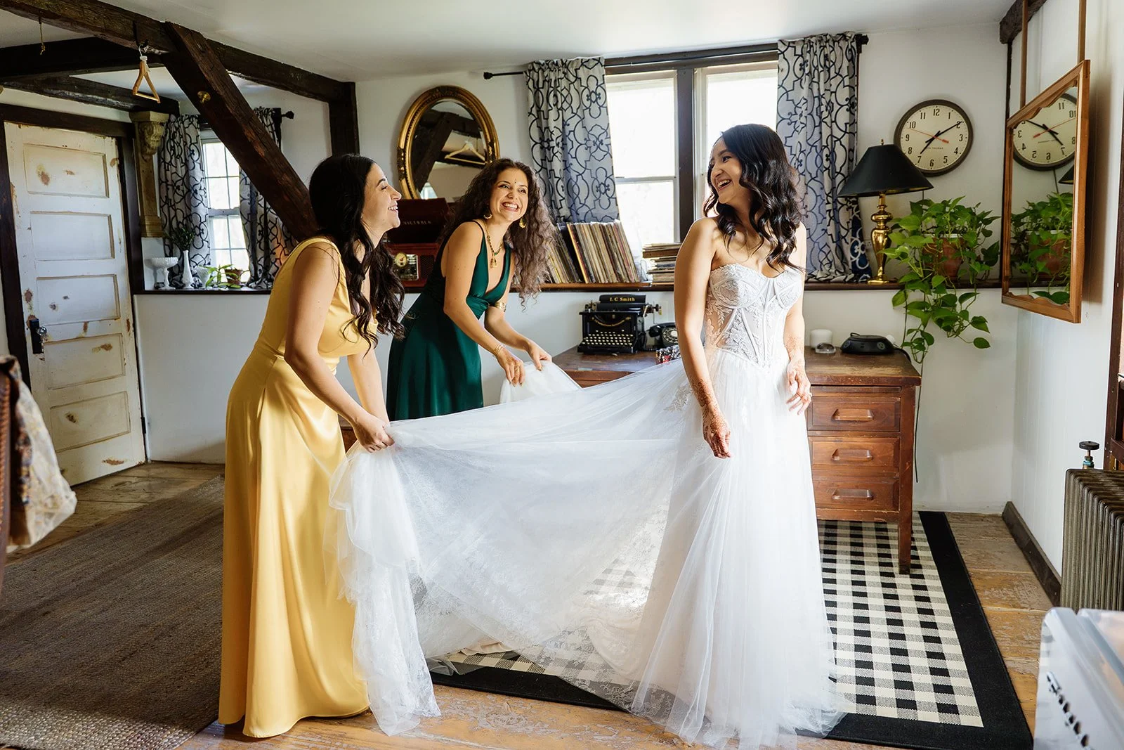 Three women, one in a white wedding dress and two in colorful dresses, are smiling and helping the bride with her dress in a cozy, rustic room with wooden beams, books, and decor.