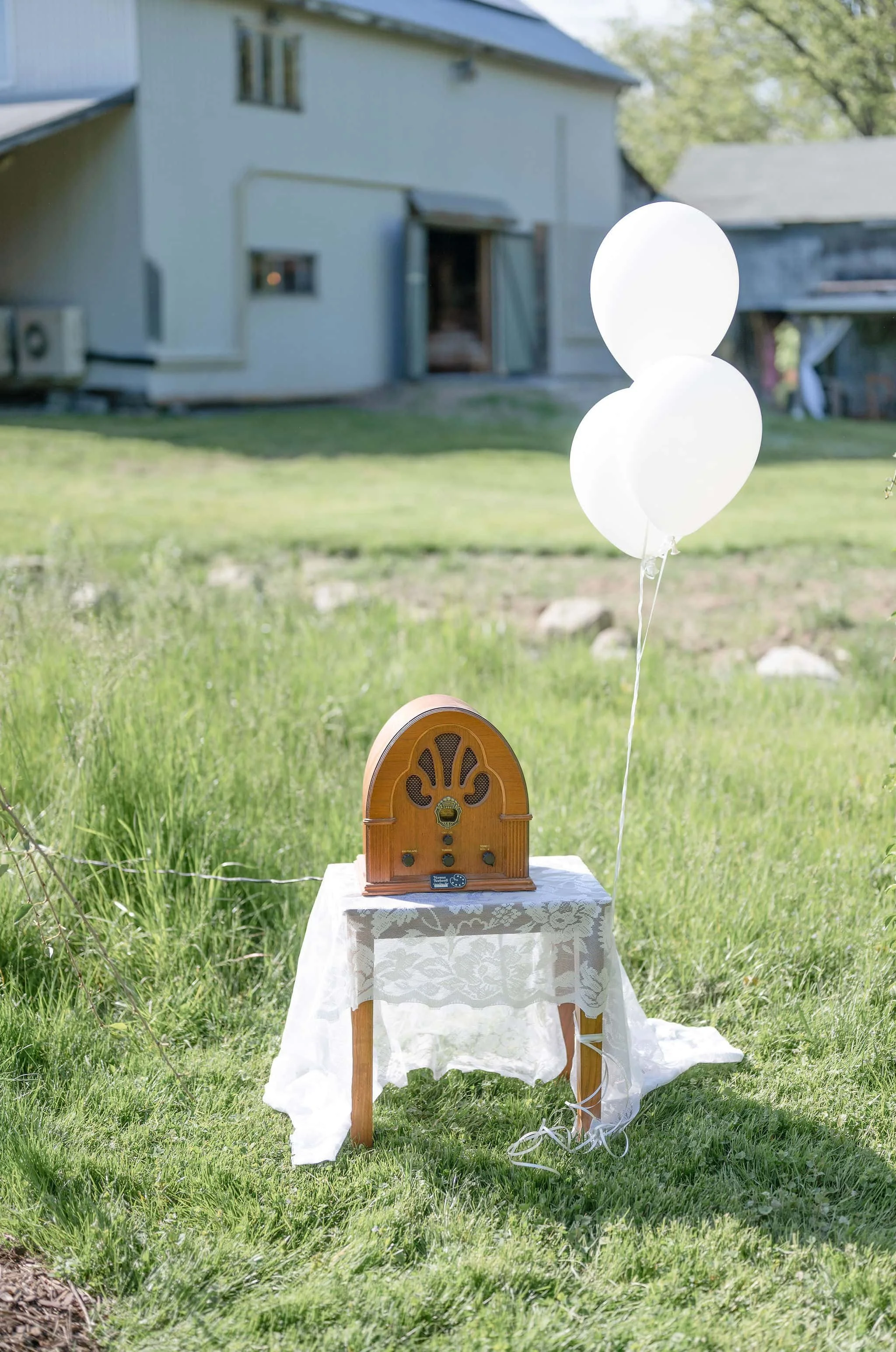 An outdoor scene with a vintage radio on a small table covered with a white lace cloth, and three white balloons tied to the table, on a grassy area with a house and trees in the background.