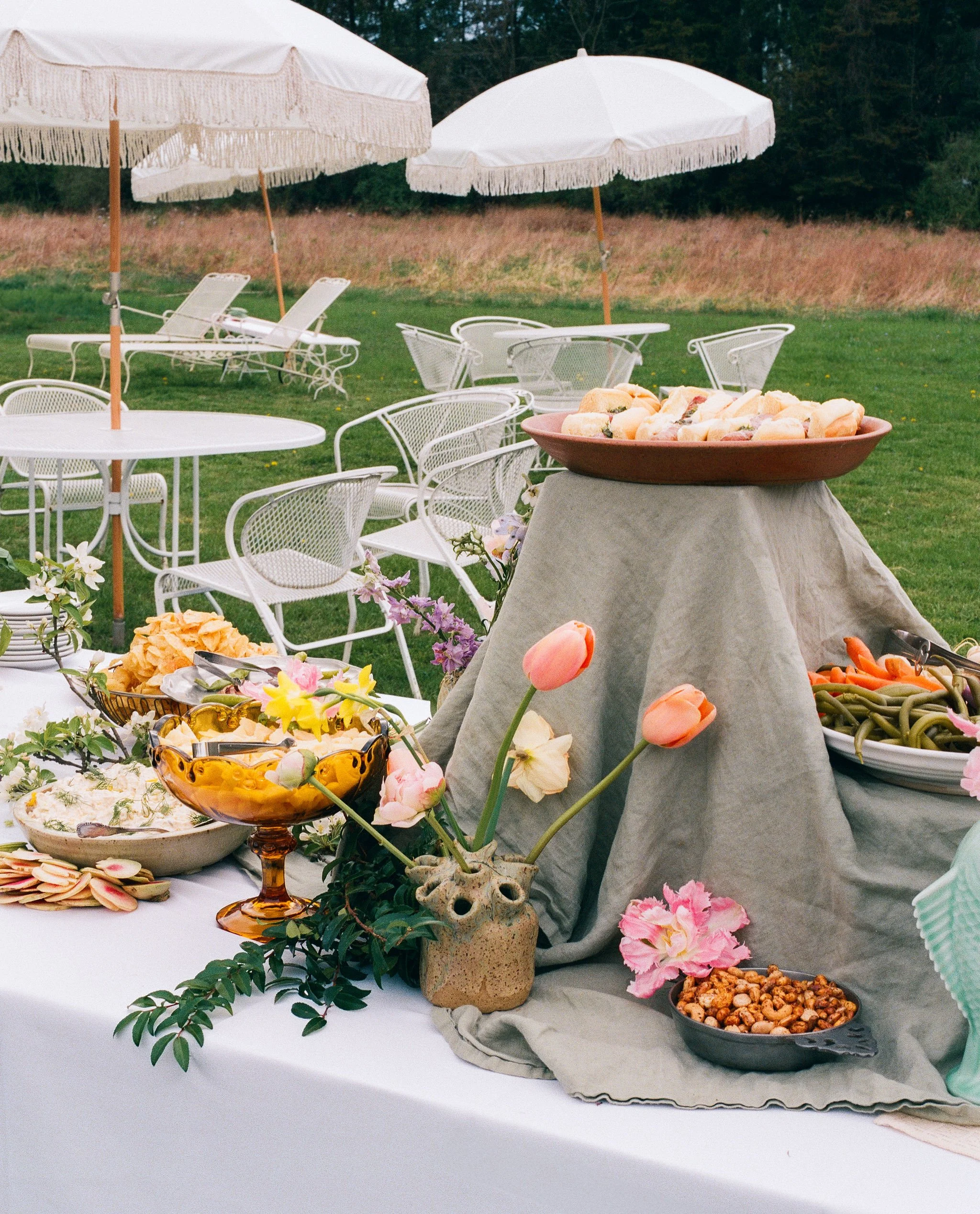 Outdoor buffet with floral decorations, assorted dishes, and pink tulips on a table with a gray cloth cover, set on a lawn with white patio furniture and umbrellas in the background.