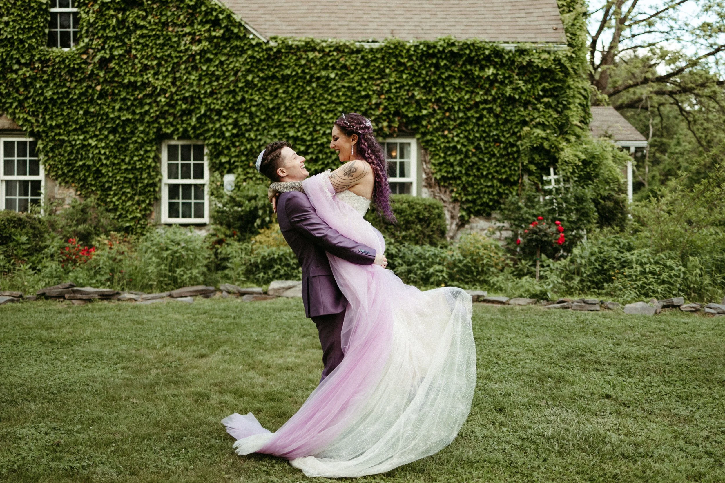 A couple in wedding attire, with the groom lifting the bride, smiling and sharing a joyful moment outdoors in a lush garden with a stone house covered in ivy in the background.