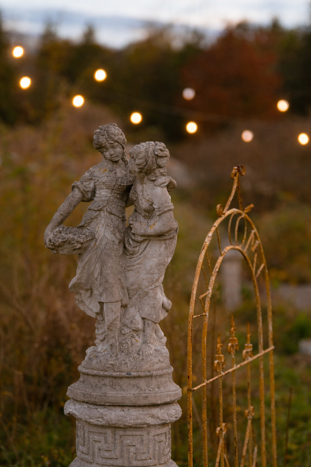 A weathered stone statue of two children, with blurred string lights in the background, and an aged, rusted metal gate nearby.