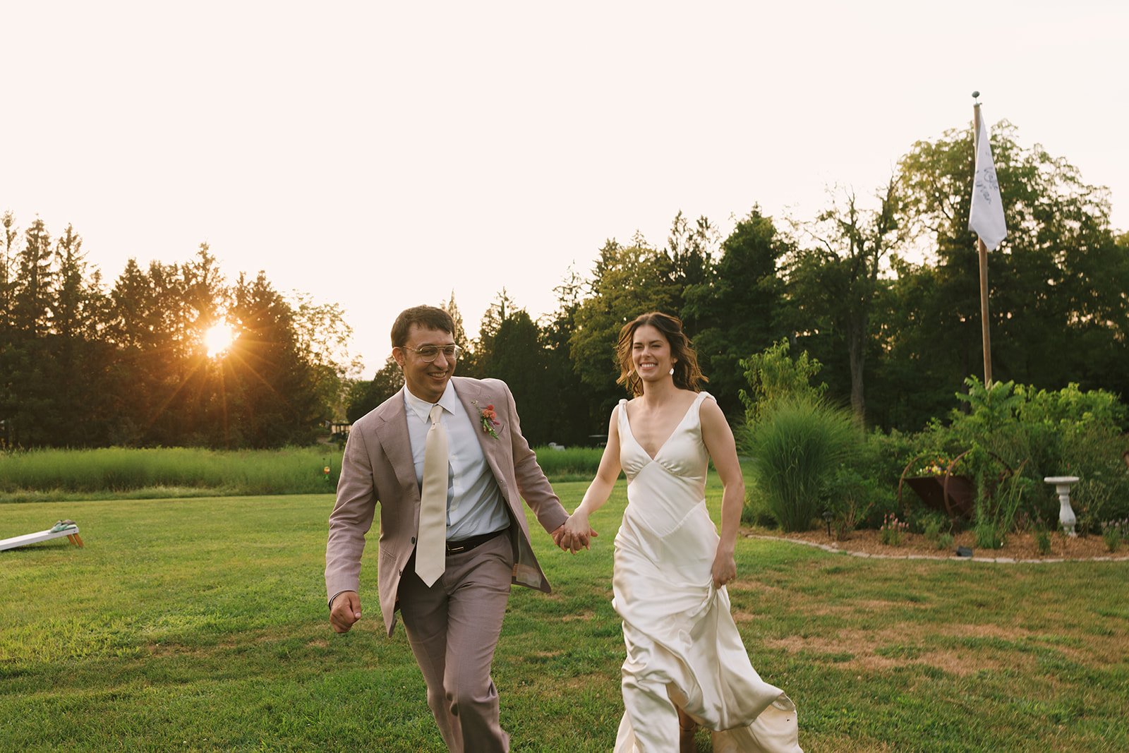 A happy couple in wedding attire holding hands and walking on a lush green lawn during sunset, with trees and a flag in the background.