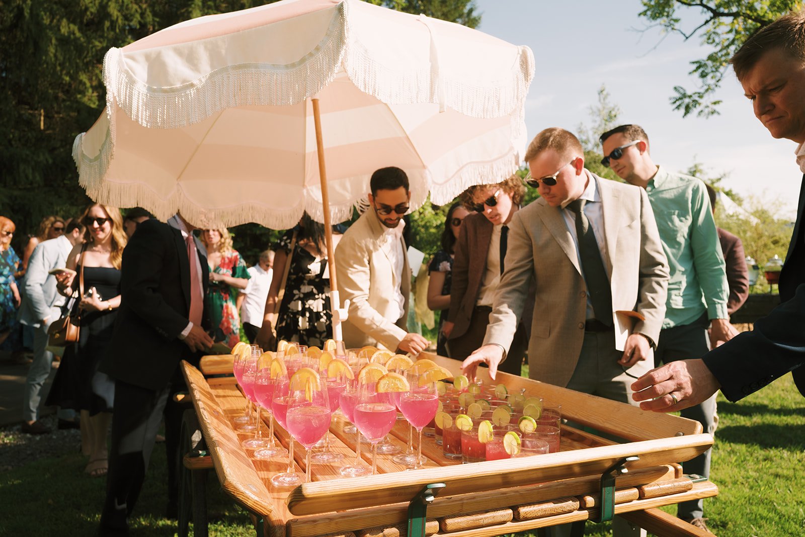 People gathered around a table outdoors, selecting drinks with pink liquids and garnished with lemon slices, under a large pink parasol.