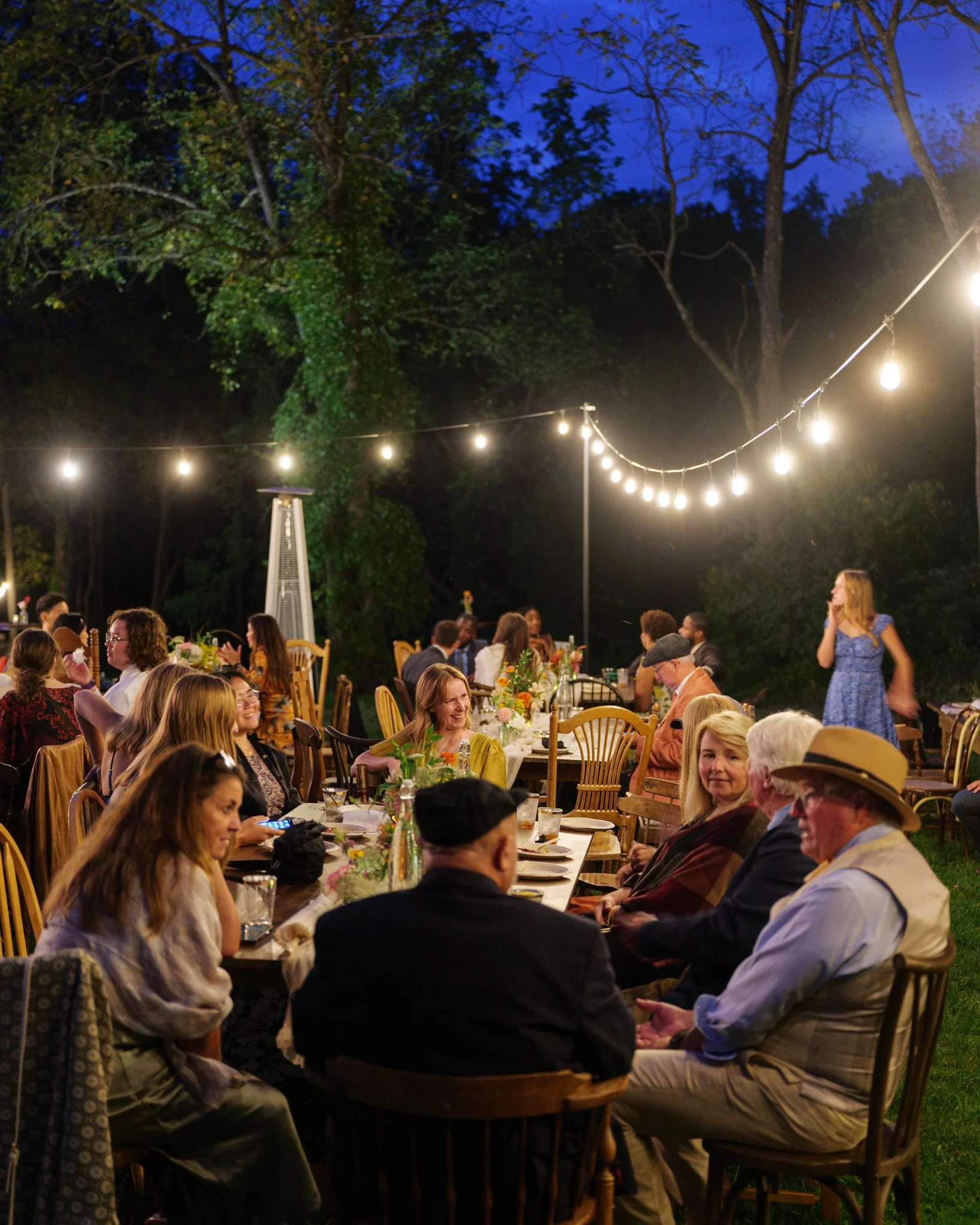 People enjoying an outdoor dinner party at night, with string lights hanging above and trees in the background.