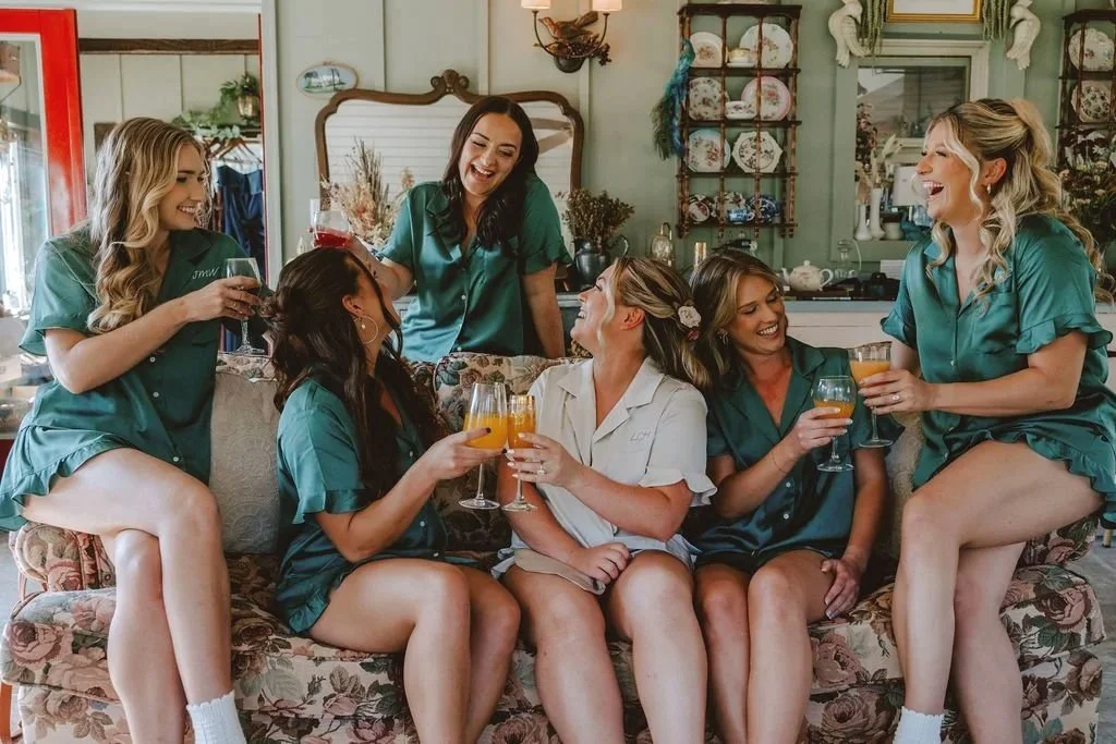 Six women in matching teal pajamas celebrating together on a floral sofa, holding drinks and smiling.