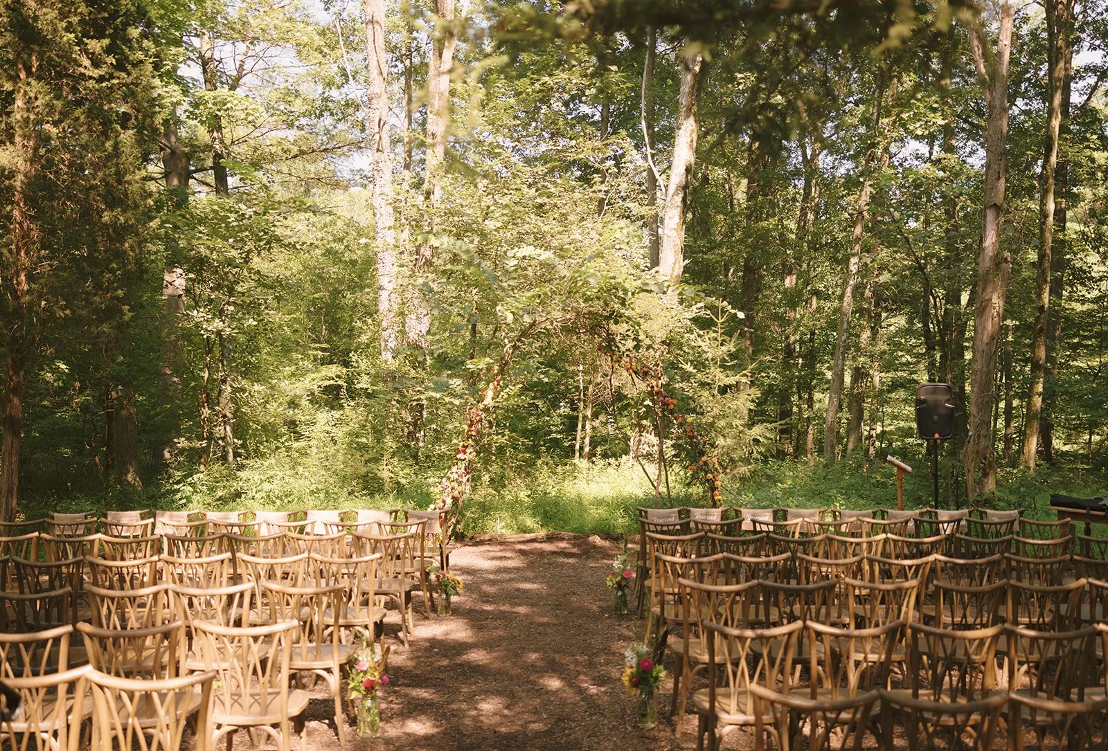 Outdoor wedding ceremony setup in a forest with rows of wooden chairs facing a floral arch and aisle decorated with flower arrangements.