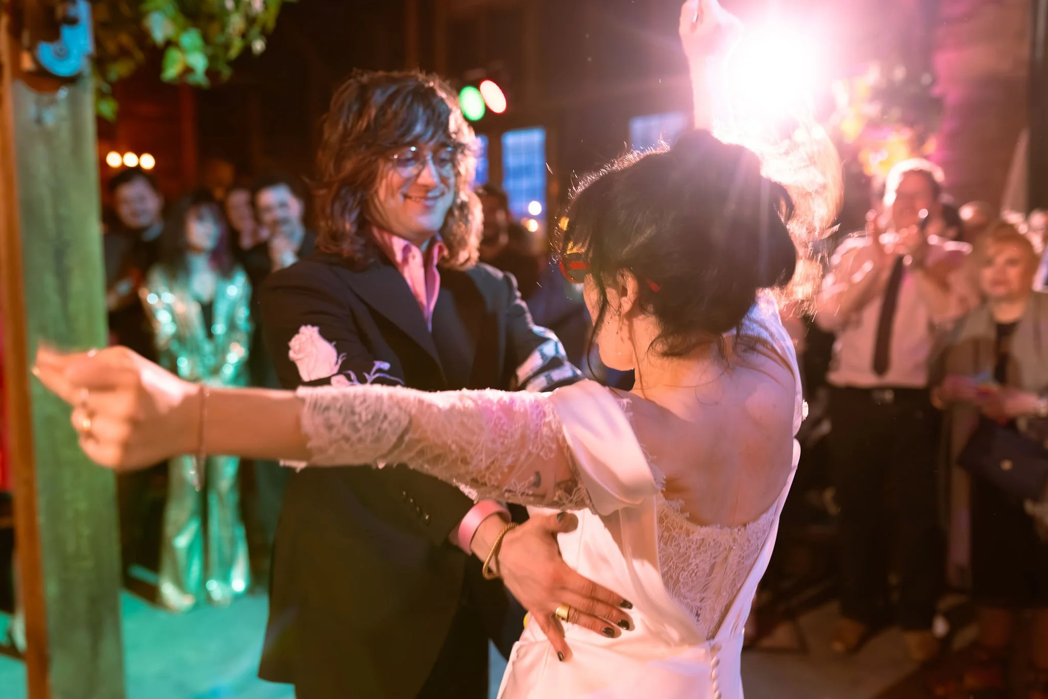A couple, a man with long hair and glasses and a woman with dark hair styled up, sharing a dance at a celebration with a crowd watching. The scene is lit with pink and purple lighting.
