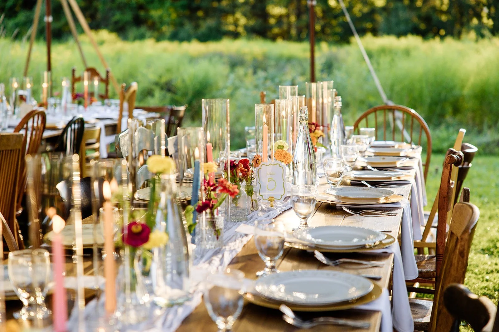 A long outdoor dining table set for a celebration with plates, glasses, and floral centerpieces, illuminated by candles, in a garden during sunset.