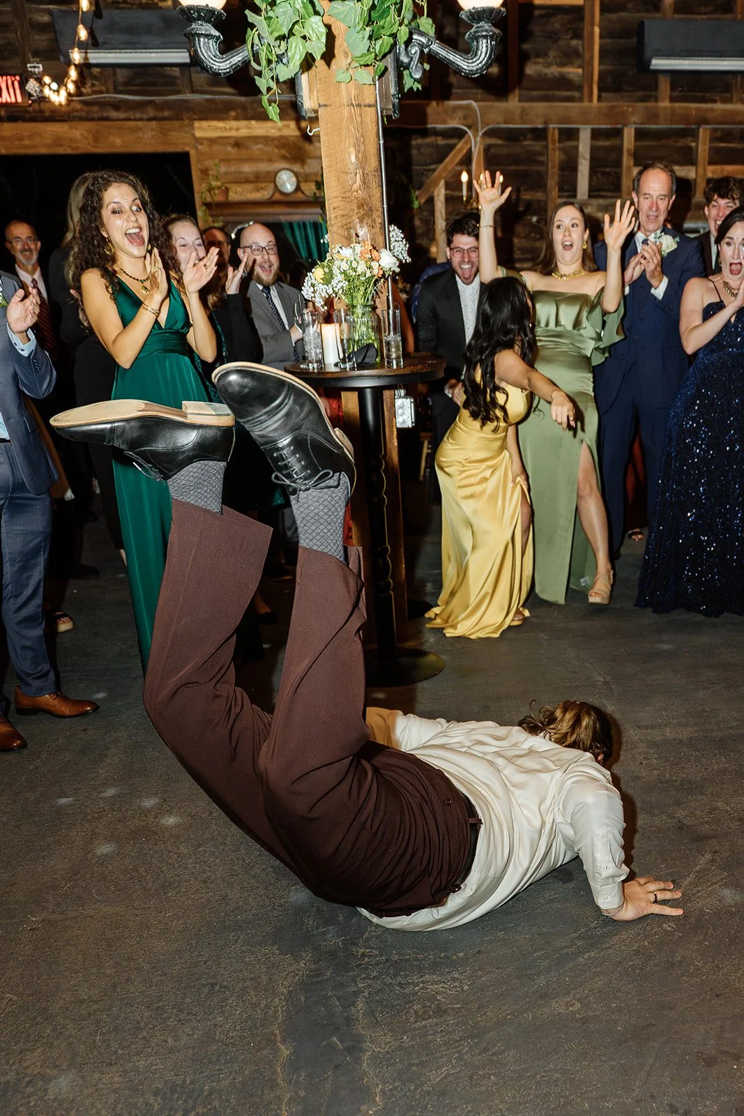 A man in a white shirt and brown pants performs a breakdance move on the floor at a party or wedding reception with smiling guests watching and cheering in the background.