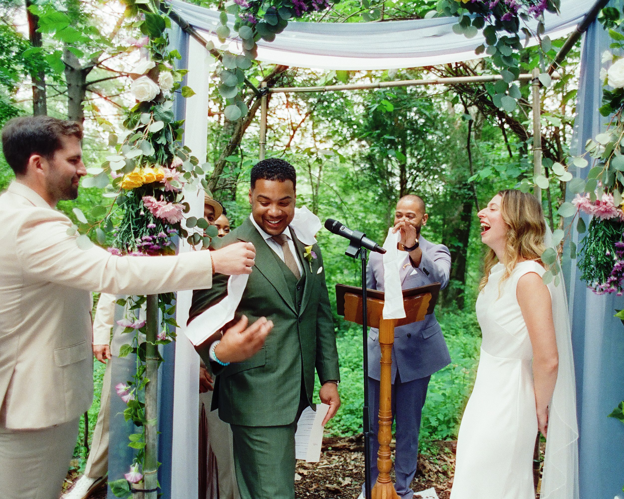 A wedding ceremony outdoors in a wooded area with greenery, where the bride and groom are standing under an arch decorated with flowers and leaves, exchanging vows with the officiant and wedding party present.