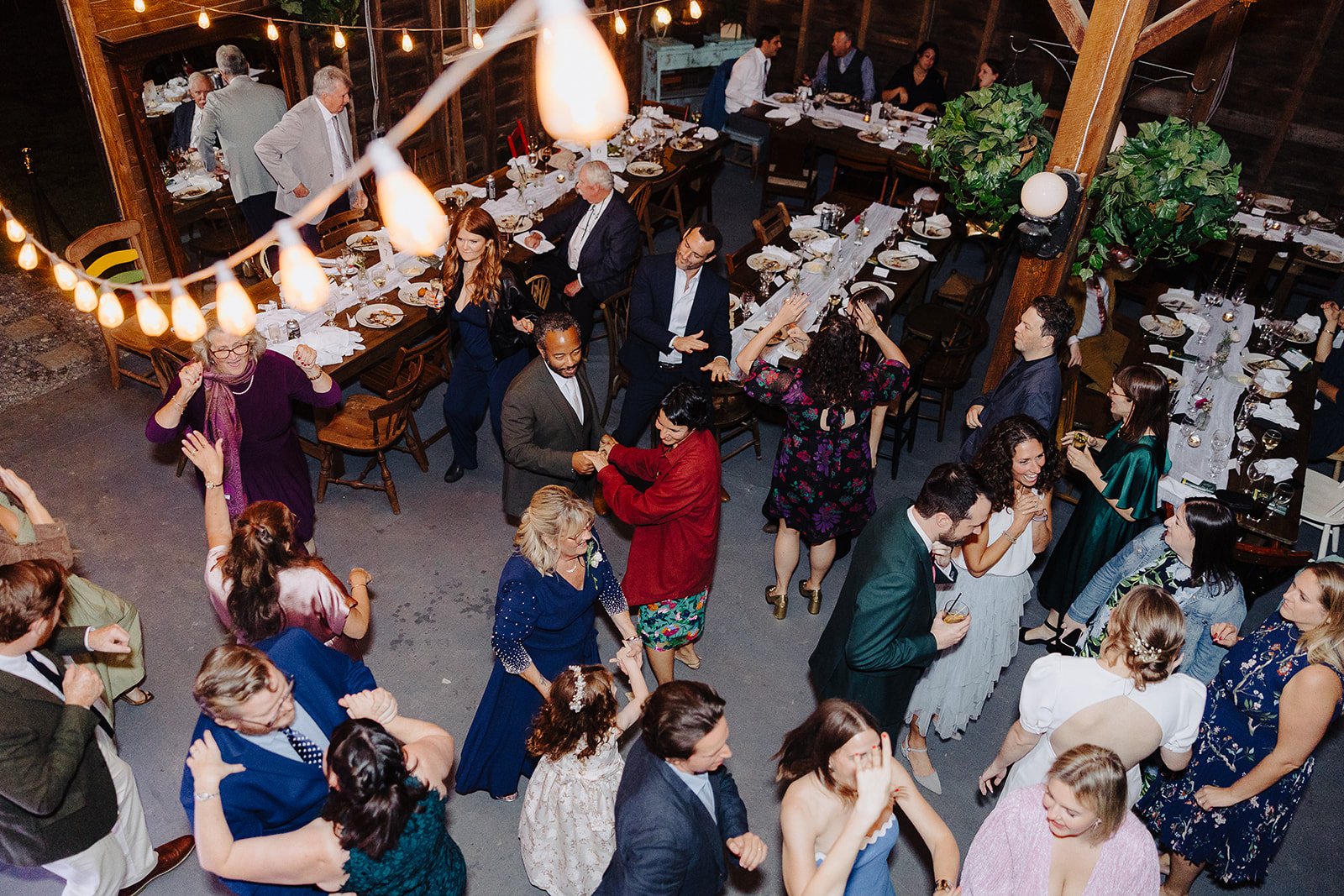 People dancing and socializing at a wedding reception or party in a rustic indoor venue with decorated tables and hanging string lights.