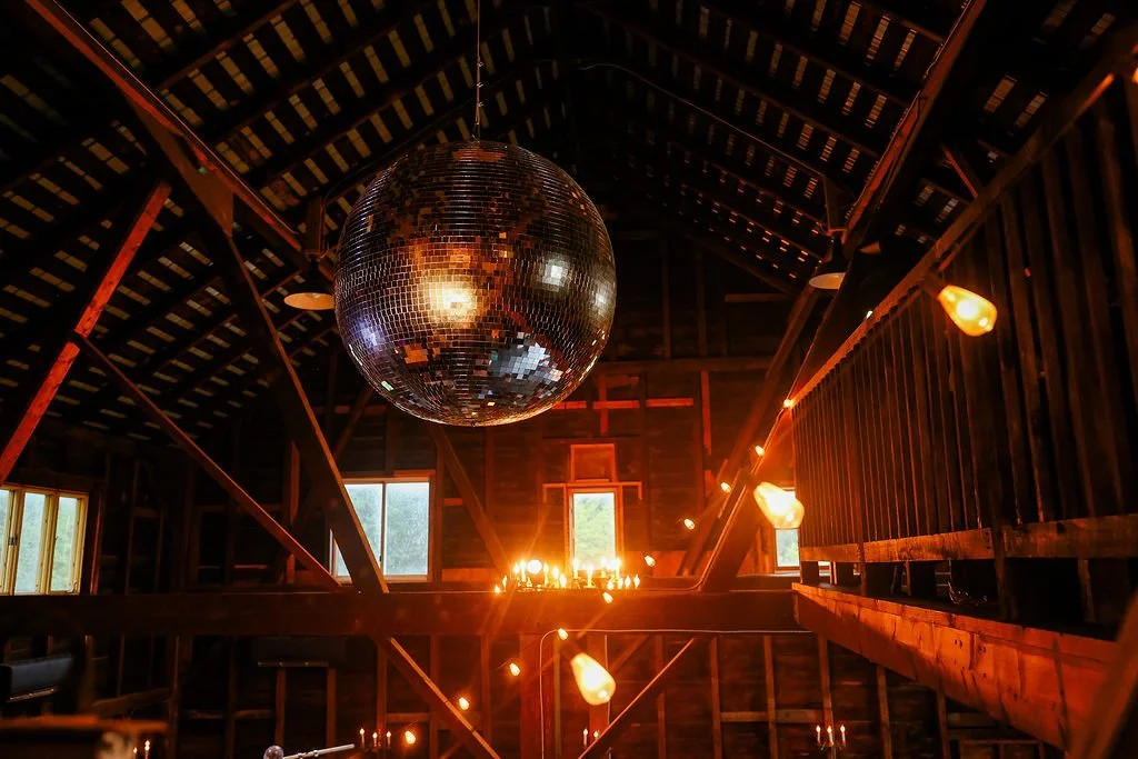 Interior of a rustic wooden barn decorated with string lights, featuring a large disco ball hanging from the ceiling and small windows letting in natural light.