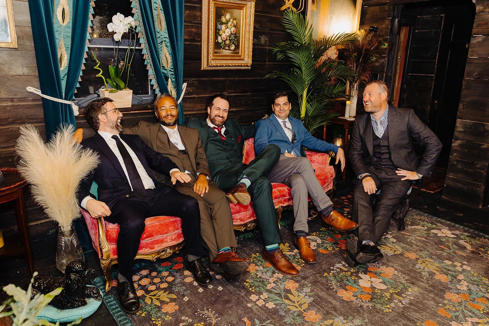 Five men dressed in suits sitting on a vintage pink and gold sofa, laughing and enjoying each other's company in a warmly decorated room with dark wooden walls, framed paintings, a large green plant, blue curtains, and a patterned floral carpet.