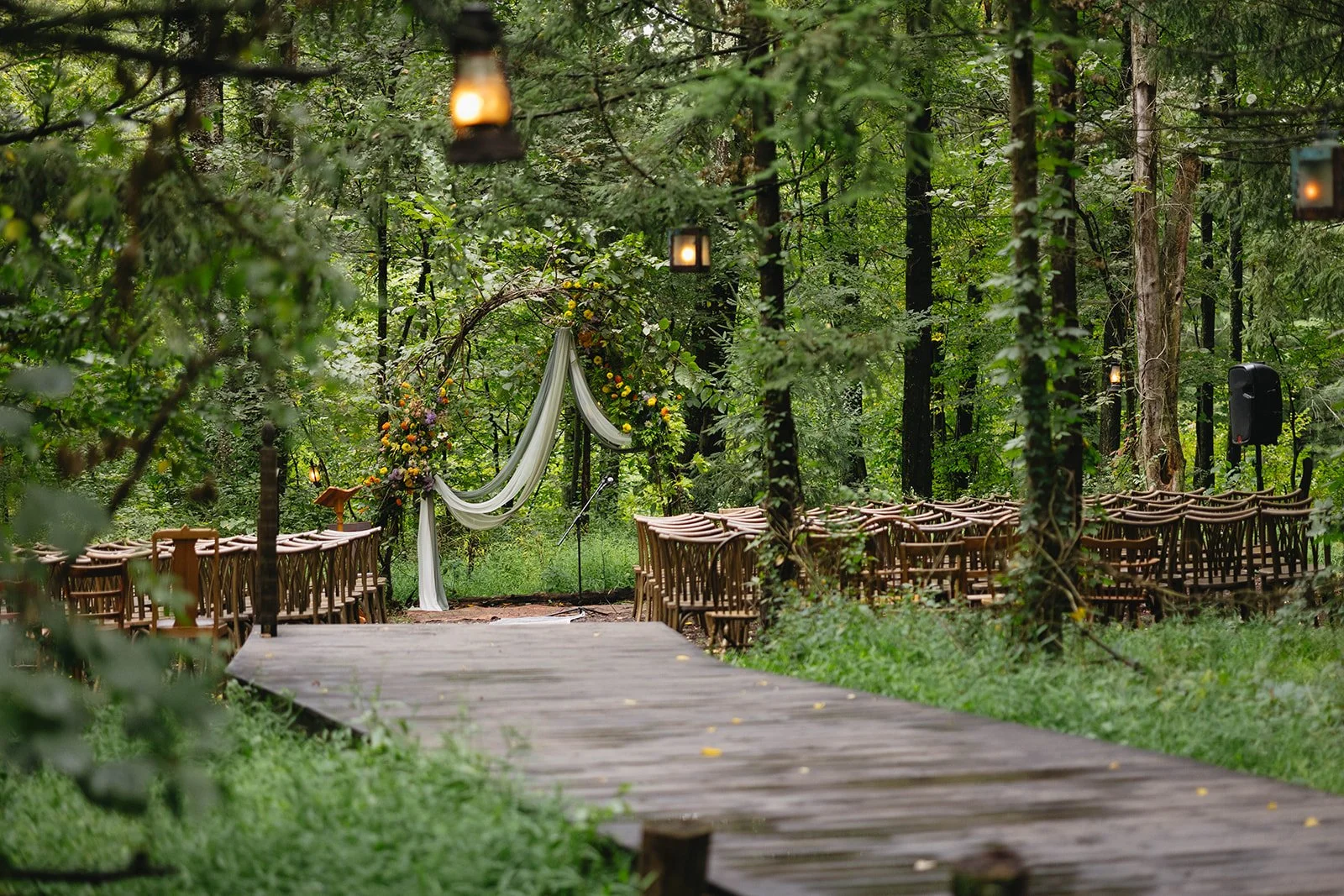 An outdoor wedding ceremony setup in a forest with wooden chairs arranged on either side of a wooden aisle, a floral arch decorated with flowers and draped fabric at the altar, hanging lanterns from trees, and a speaker in the background.