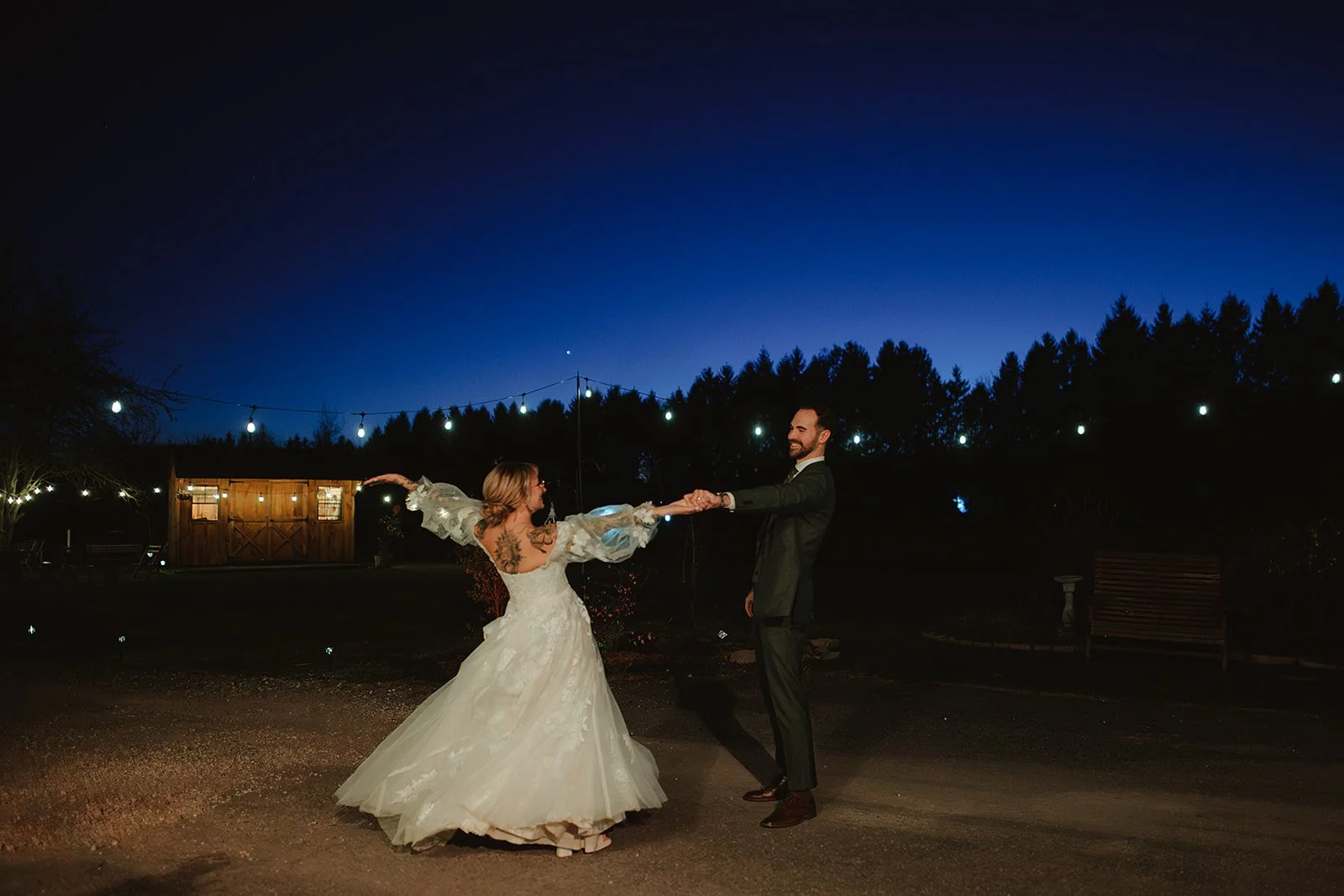 A bride and groom dancing outdoors at night with string lights overhead and a wooden barn in the background.