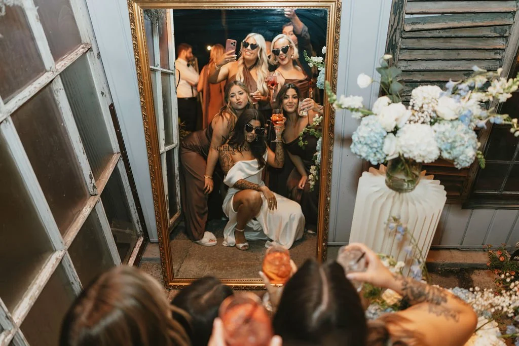 A group of women taking a selfie in front of a large mirror at a celebration or party, with some women holding glasses of drinks and posing playfully. The scene is decorated with flowers in a vase and a rustic setting.