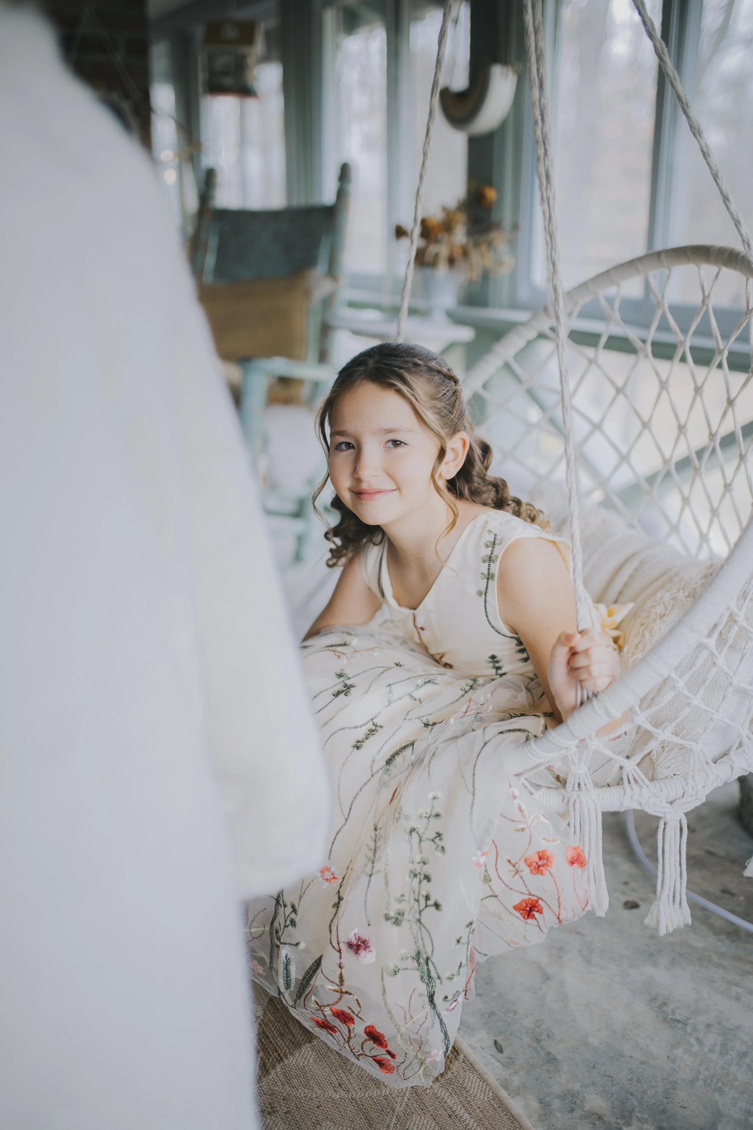A young girl with curly brown hair in a white embroidered dress is sitting in a hanging chair inside a sunlit room, smiling at the camera.
