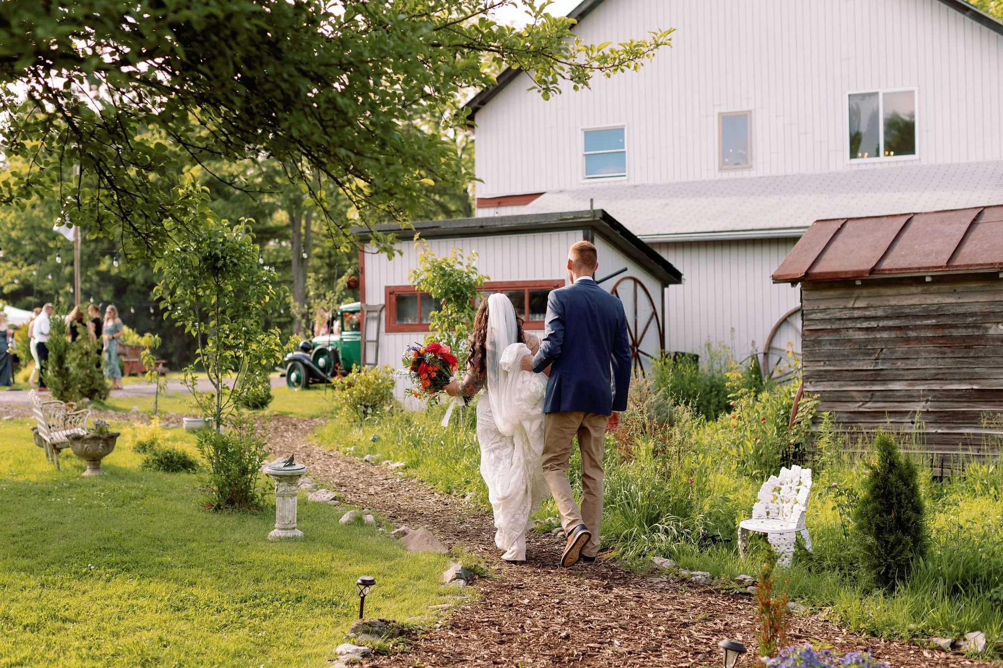 A bride and groom walking arm-in-arm on a garden path, with the bride holding a bouquet of flowers. They are near a white barn with windows, surrounded by greenery and garden decorations.