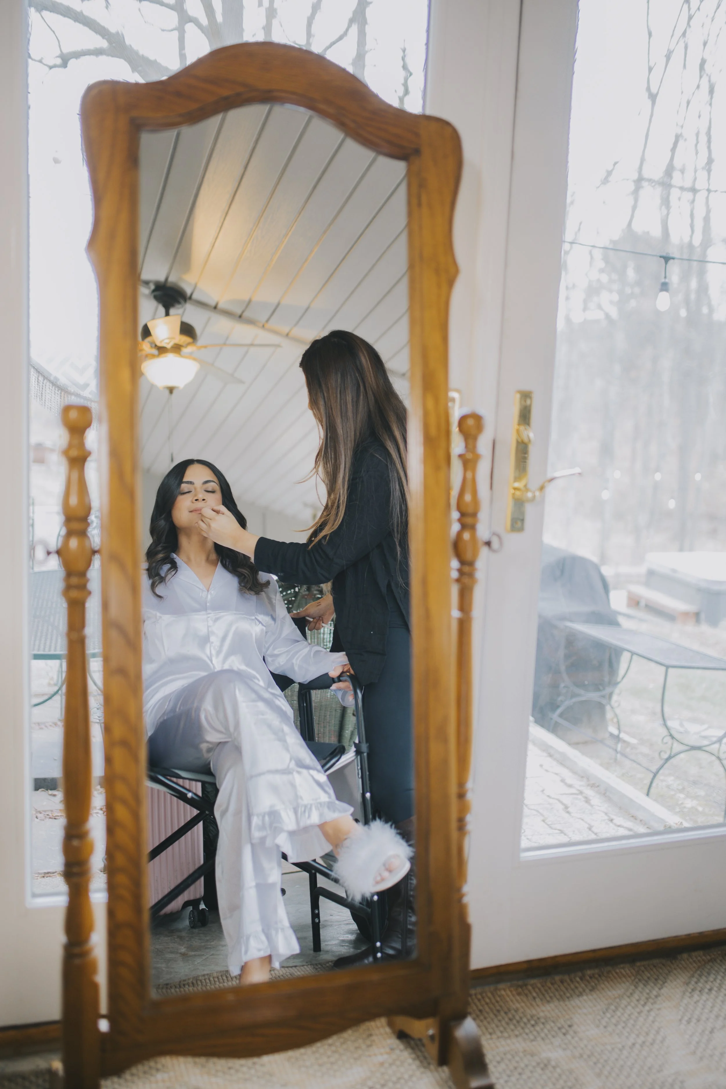 A woman in a white satin robe getting makeup applied while sitting in a chair, reflected in a tall wooden mirror, with a large window showing an outdoor porch in the background.