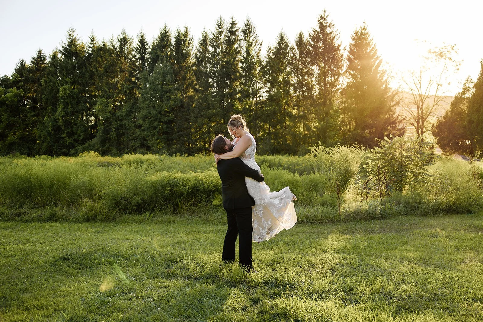 A couple in wedding attire sharing a loving embrace in a green field at sunset with tall trees in the background.