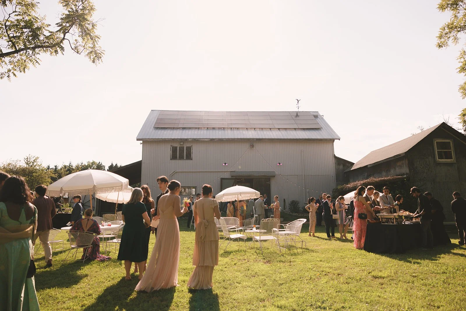 Group of people gathered outdoors near barn, some standing, some sitting under umbrellas, at a social event on a grassy area with sunny weather.
