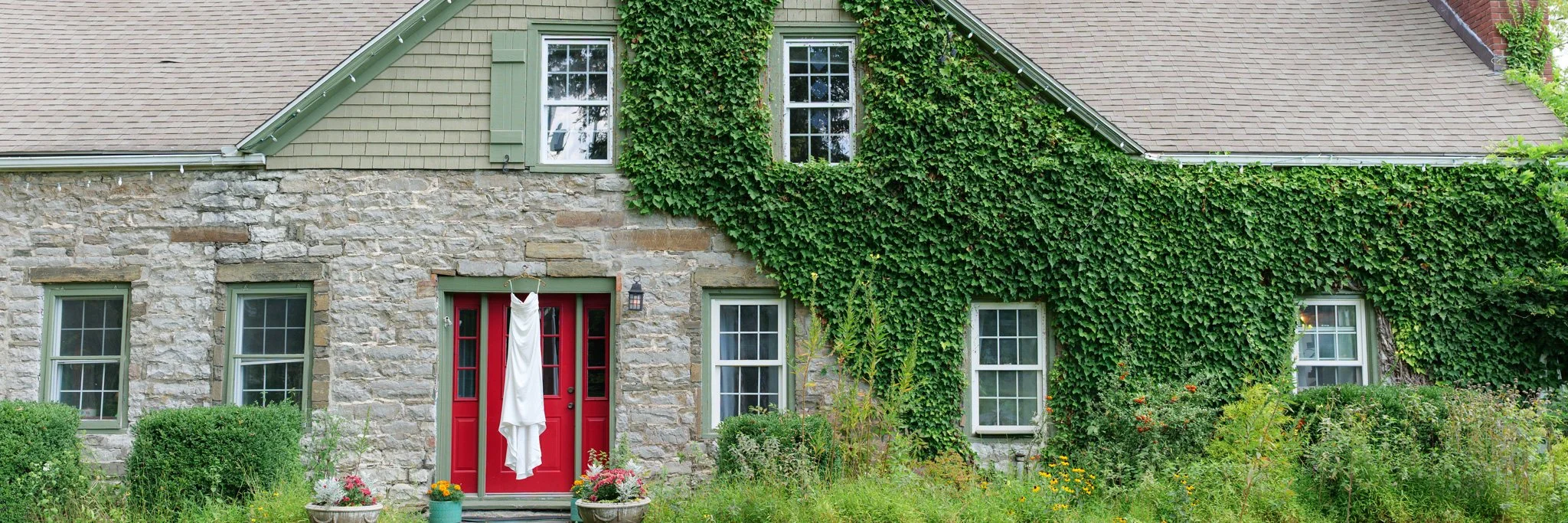 A white bridal gowns hangs in front of an ivy-covered stone house in the Hudson Valley at Black Walnut Farm