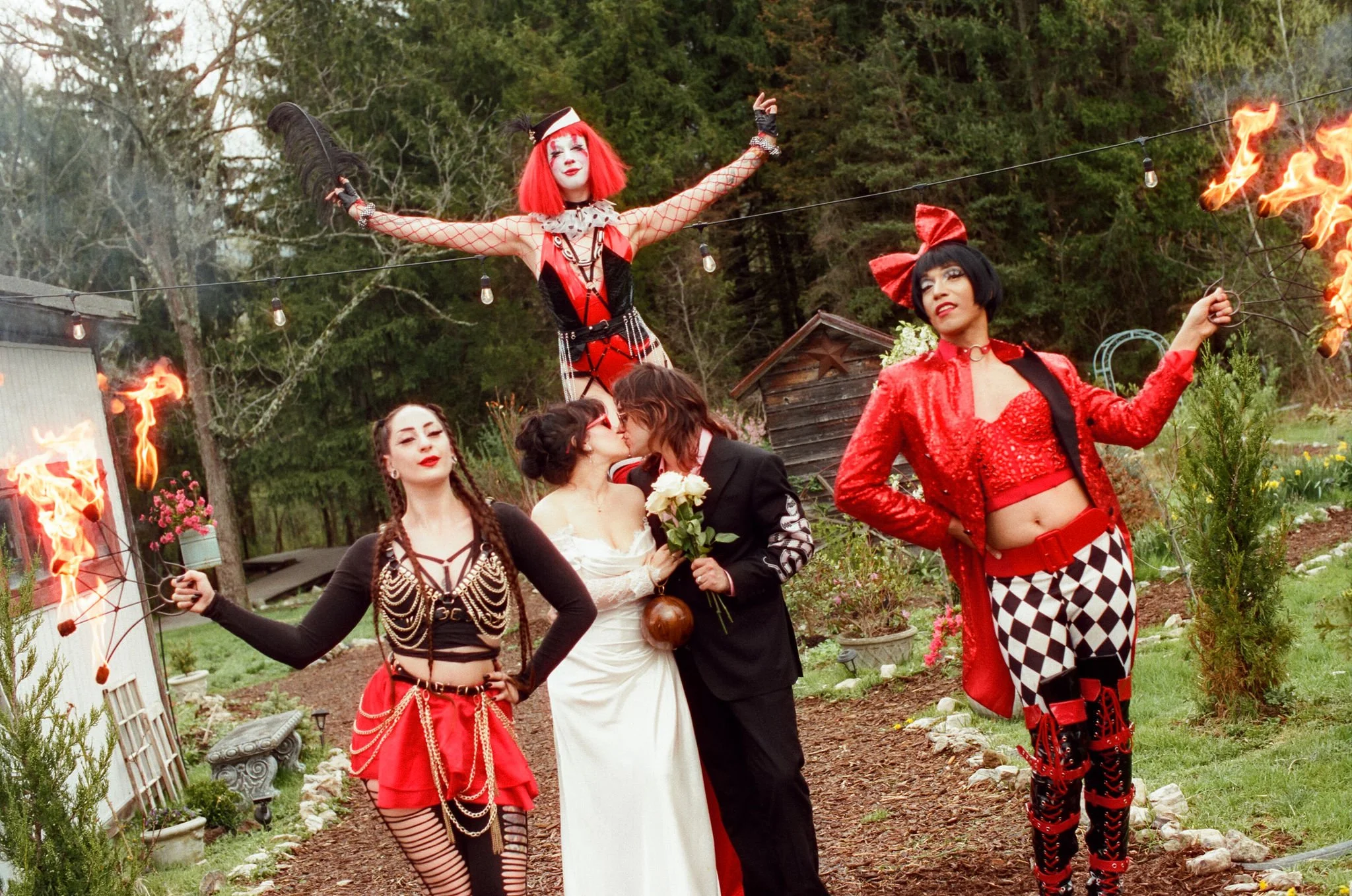 A group of people dressed in circus-inspired costumes during a celebration, with two women kissing, surrounded by performers with flaming props.