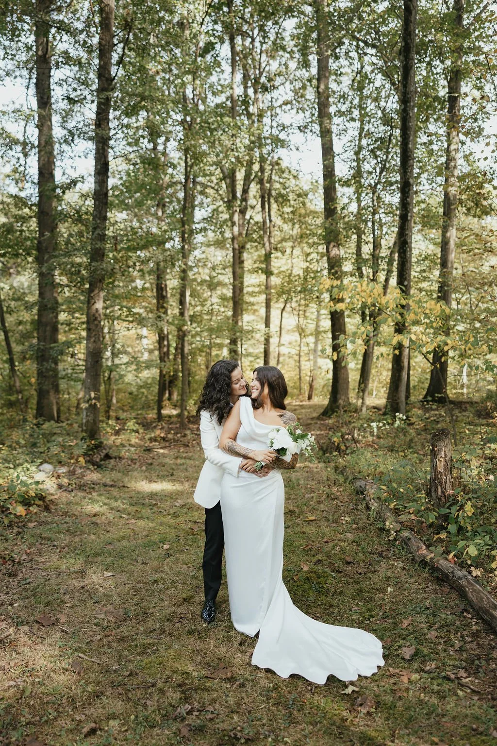 Two women dressed in wedding attire stand close together in a forest, smiling and holding a bouquet of white flowers, surrounded by tall trees and autumn foliage.