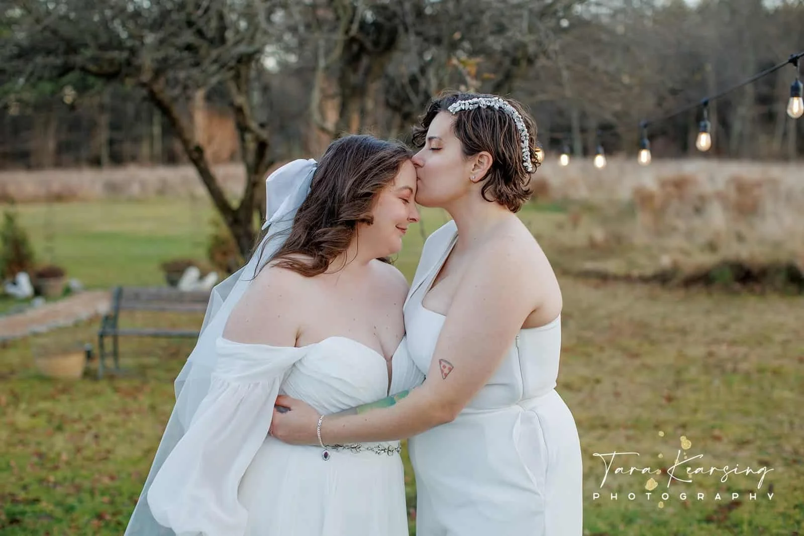 Two women in white dresses sharing a kiss on the forehead outdoors, with string lights and trees in the background, during dusk or evening.
