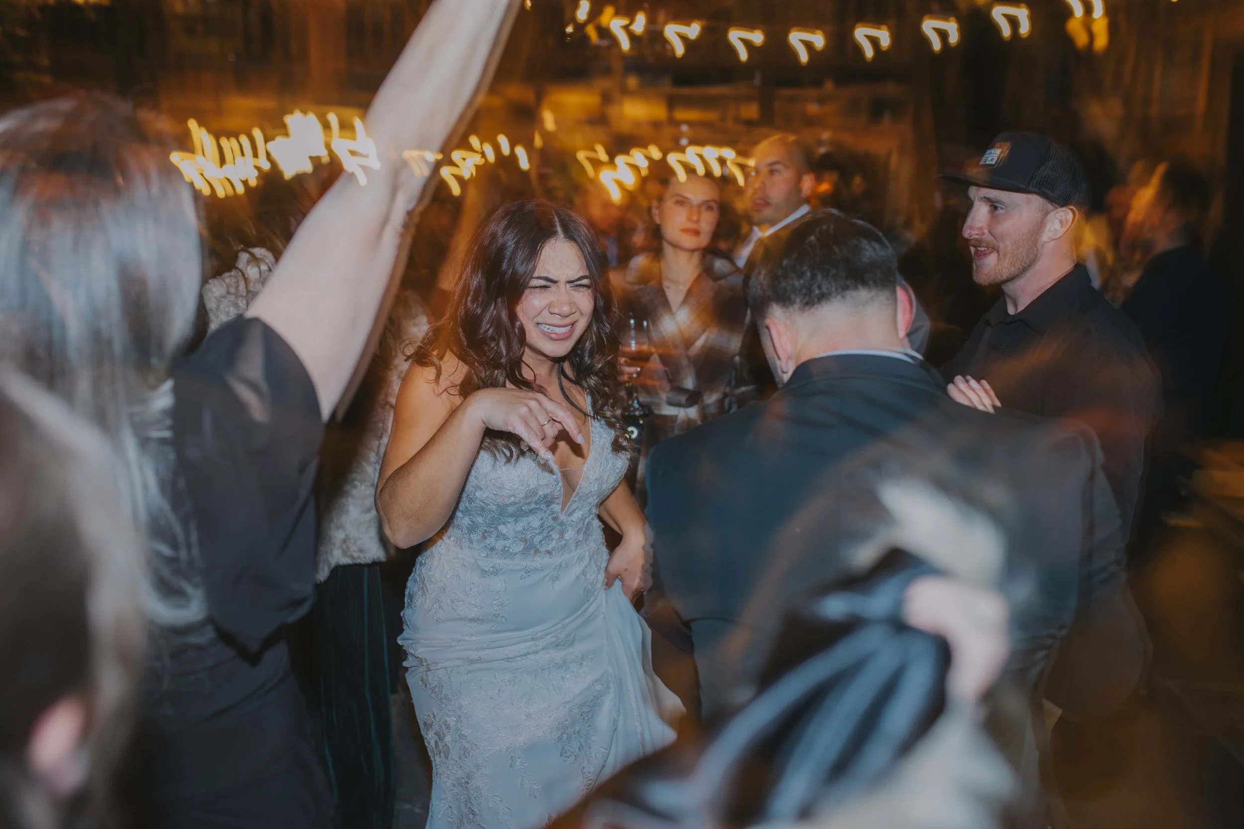 A woman in a wedding dress dancing with friends at a party, with warm string lights hanging overhead.