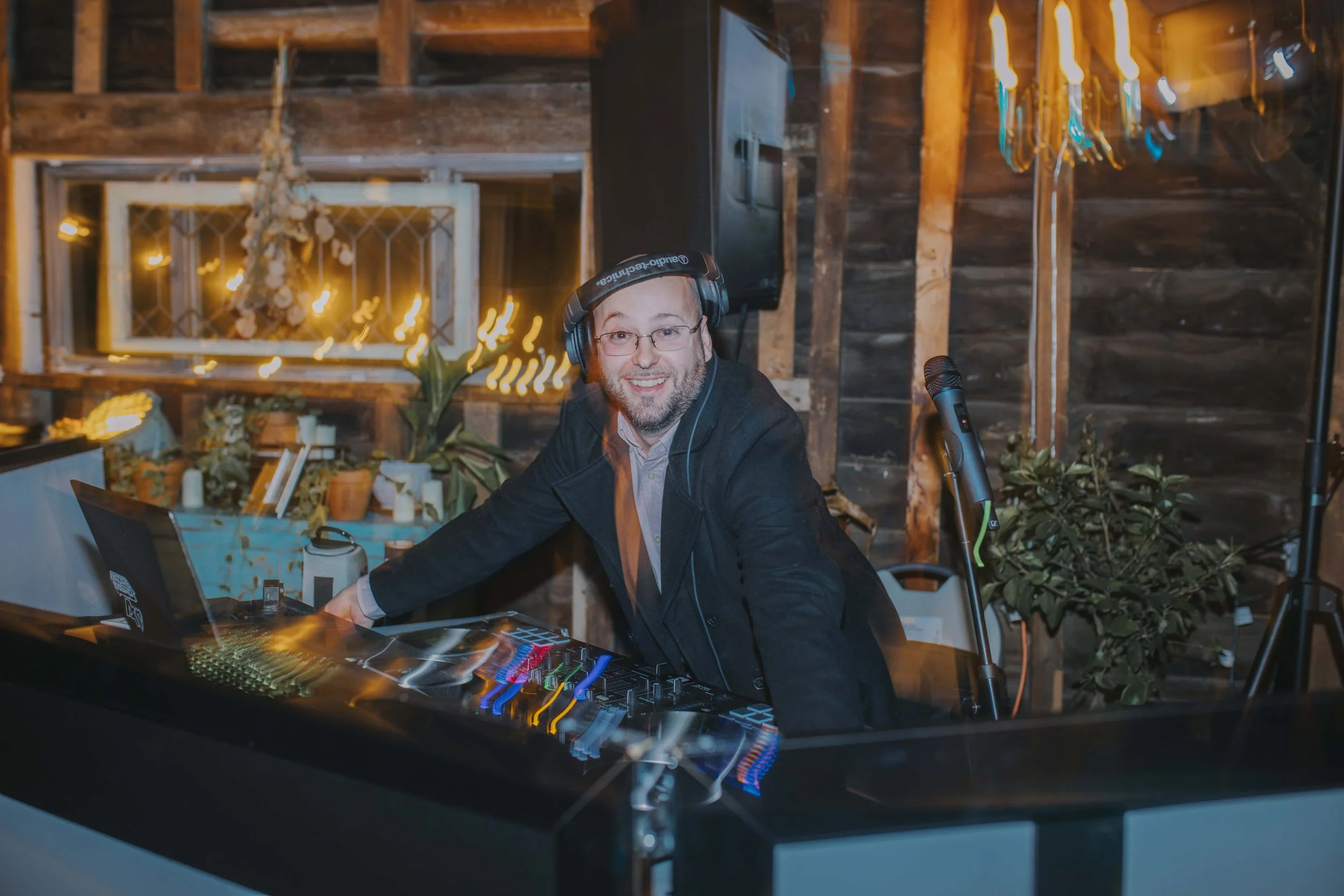 A man DJ wearing headphones, smiling, and operating a DJ mixer and laptop in a warmly lit rustic setting.