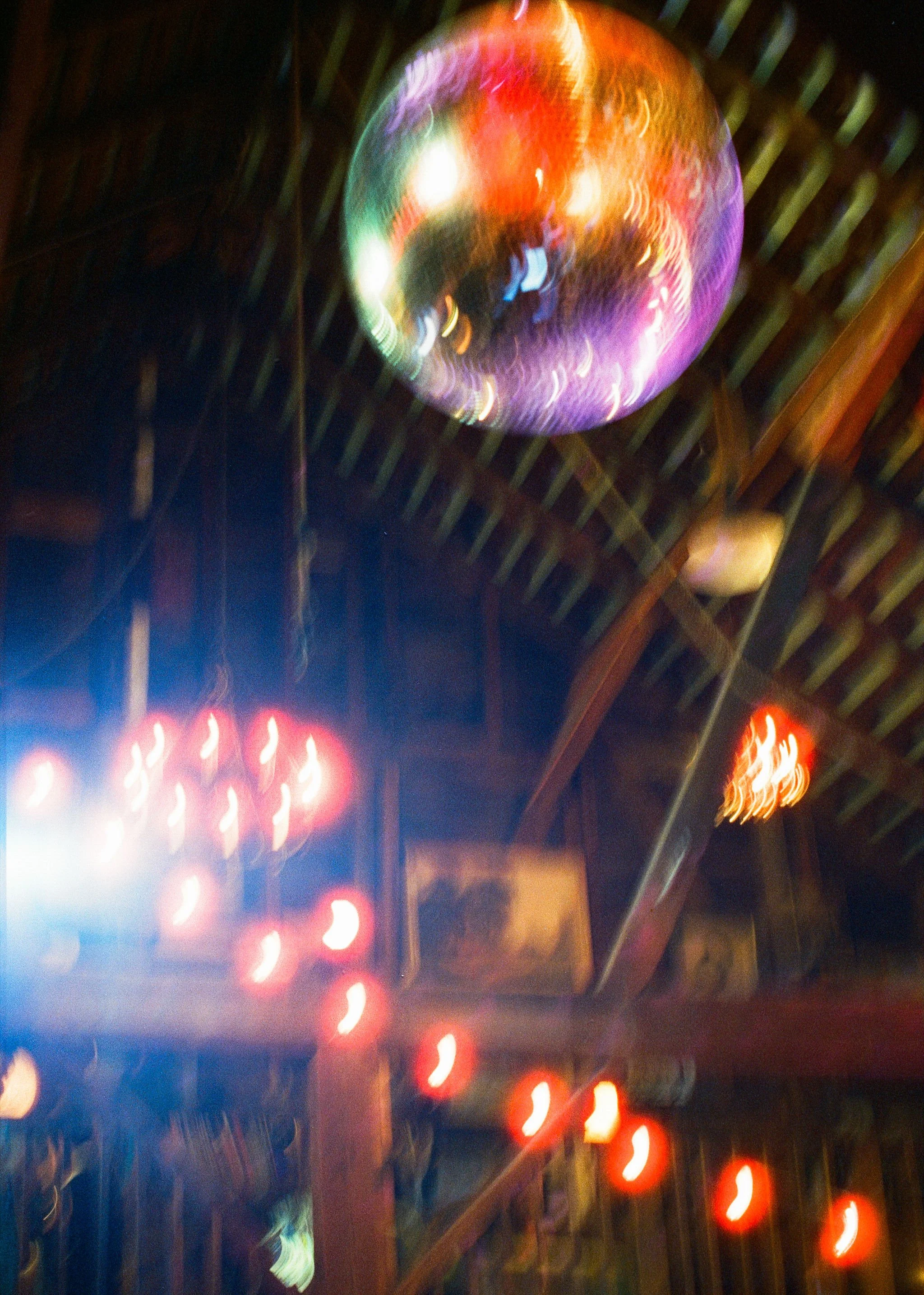 A colorful, reflective disco ball hangs from a ceiling with wooden beams. The scene features blurred, glowing red string lights and a neon sign in the background, suggesting a lively, indoor party or bar setting.