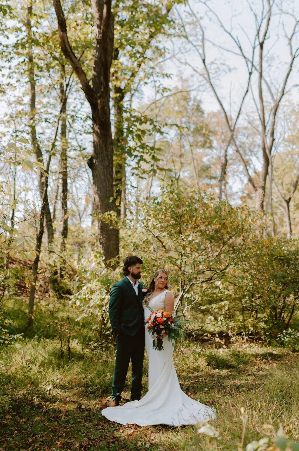 A bride in a long white wedding dress holding a colorful bouquet stands next to a groom in a dark suit, outdoors in a wooded area with trees and autumn leaves.