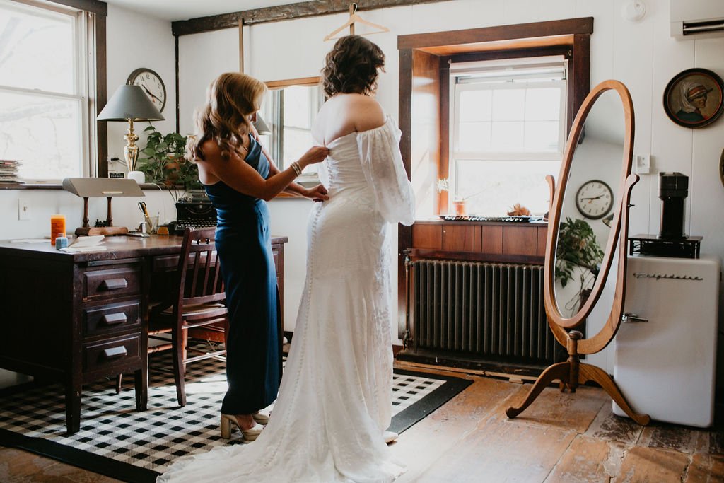 A bride getting zipped into her gown by her mother in front of a full length mirror