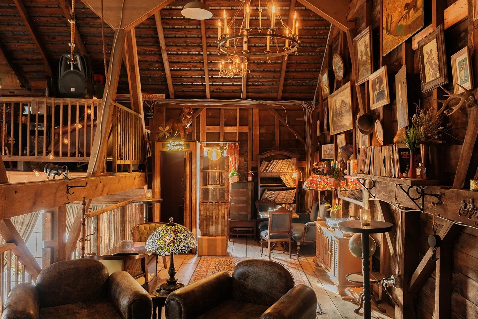 A cozy rustic living room with wooden walls and ceiling, vintage furniture, a chandelier, various framed pictures and paintings on the wall, bookshelves, a guitar, and decorative lamps.