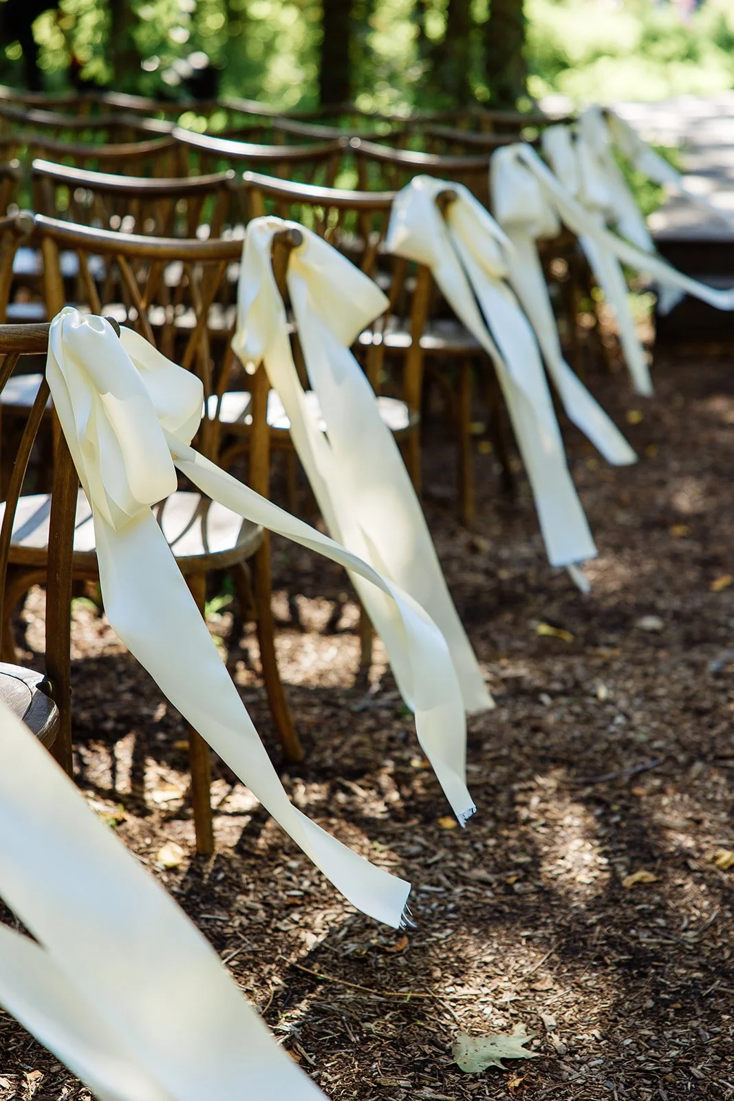 Wooden chairs decorated with white ribbons, arranged outdoors on a dirt ground for a wedding ceremony.