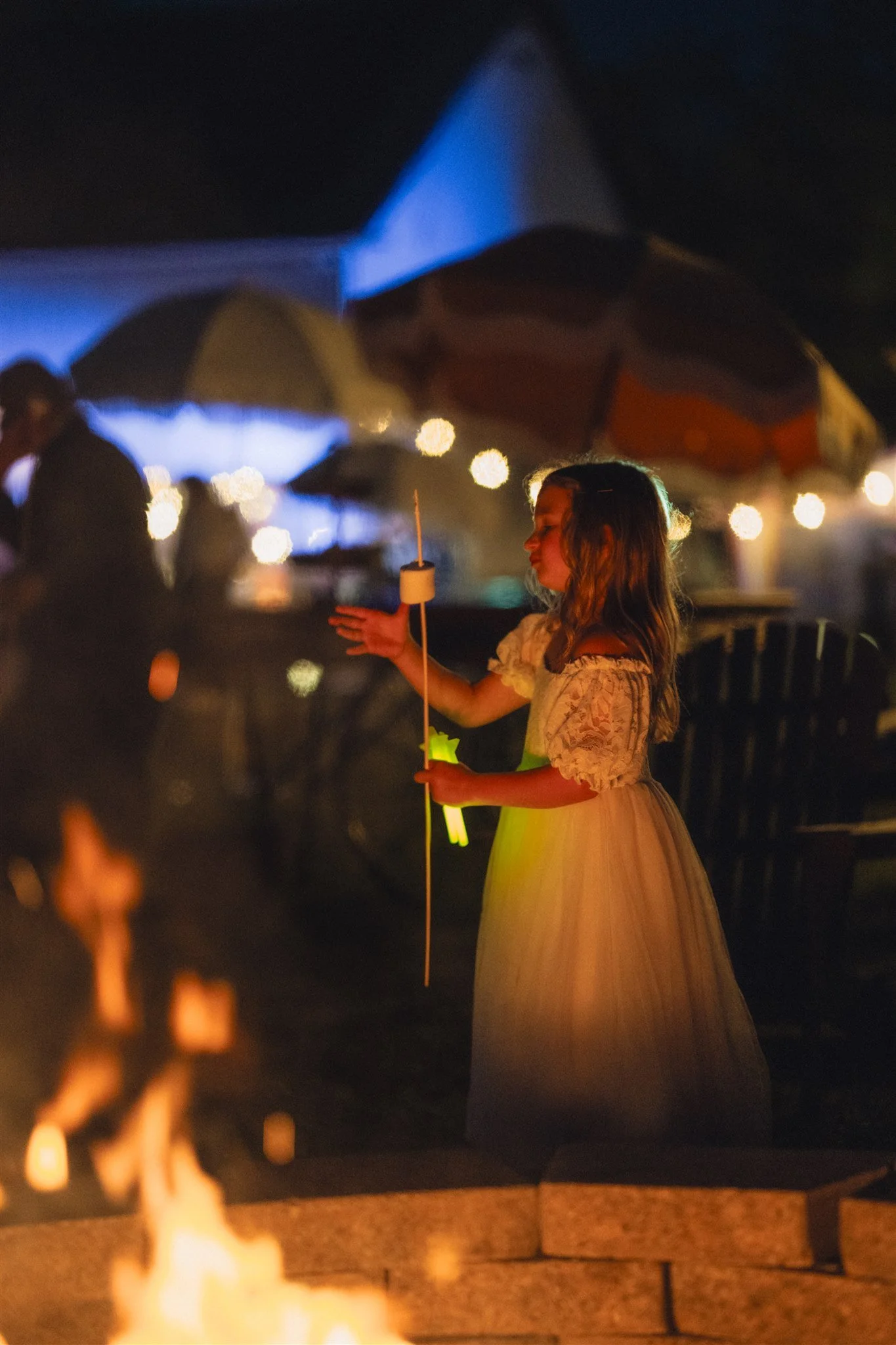 A young girl in a white dress holding a glow stick and marshmallow skewer, standing by an outdoor fire pit at night with illuminated tents and string lights in the background.