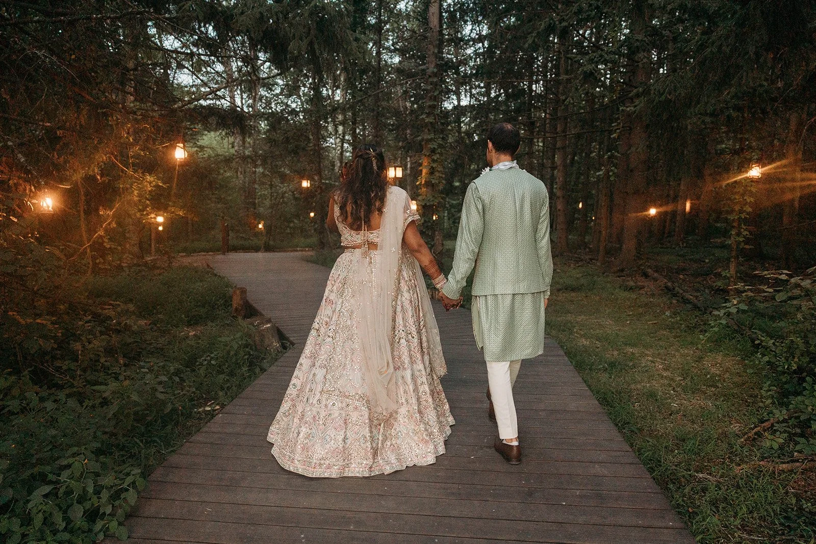Couple in traditional Indian wedding attire holding hands, walking on a wooden pathway through a forested area illuminated by hanging lanterns during dusk or evening.