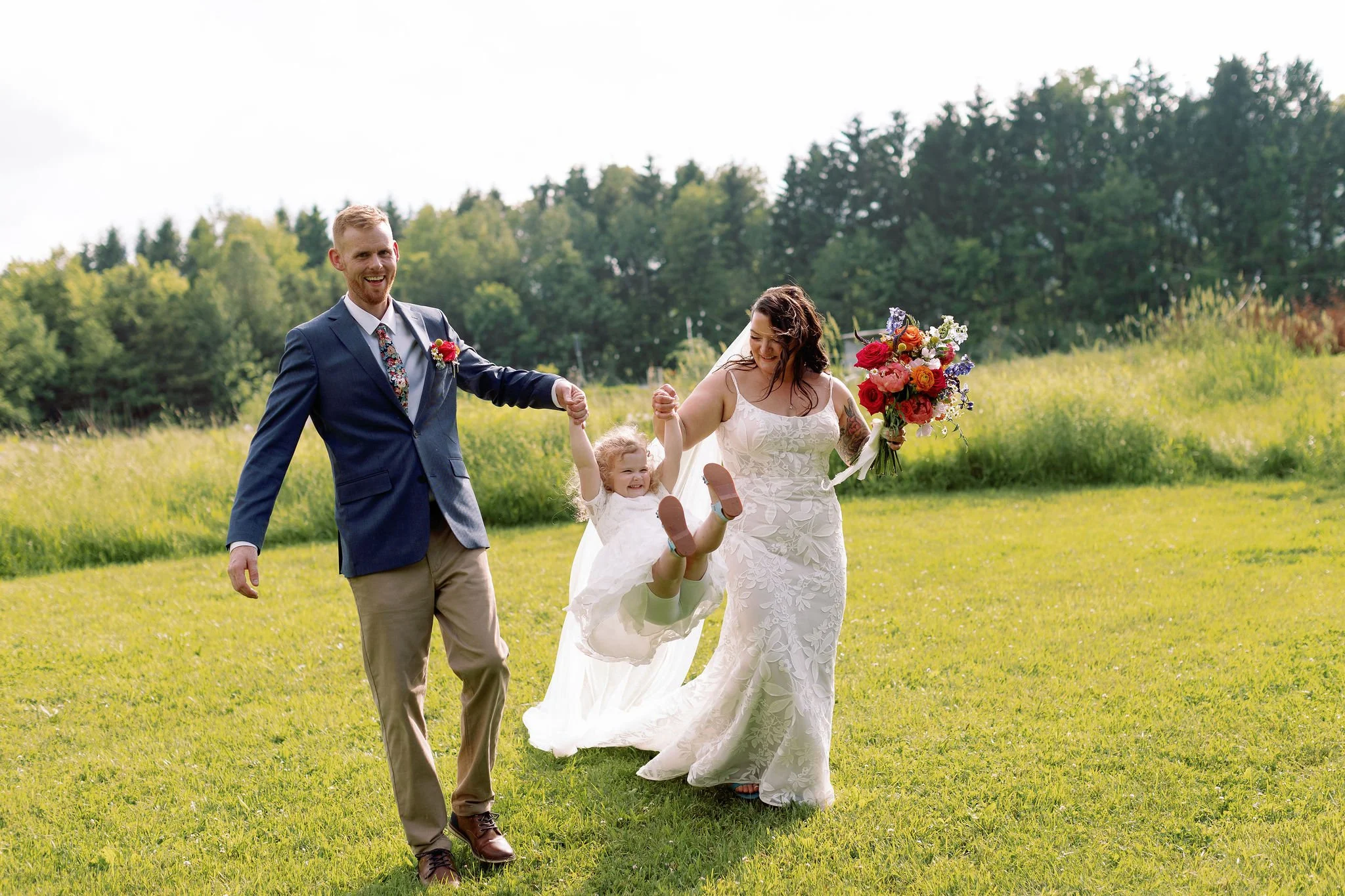 A bride and groom holding hands with a young girl between them outdoors on a grassy field, the bride holding a bouquet, all smiling happily.