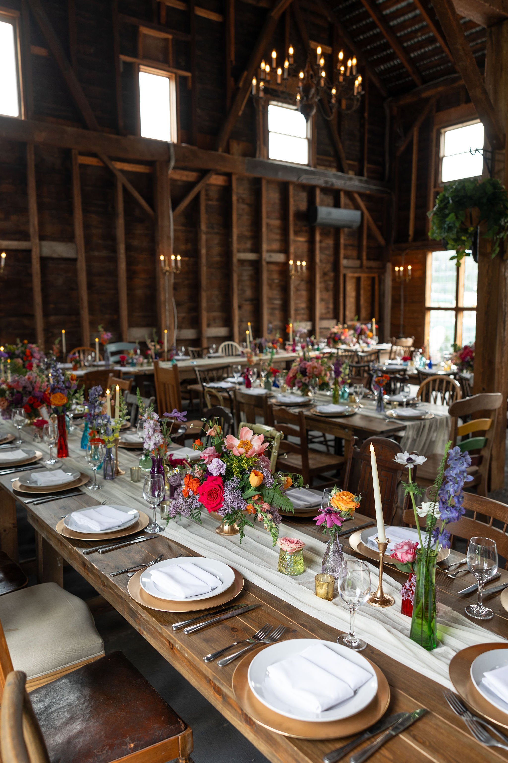 Long wooden banquet table decorated with colorful flower arrangements and gold candle holders, set for a formal event, inside a rustic wooden barn with high ceilings and chandeliers.
