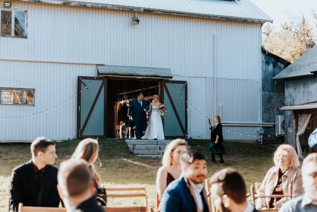 Couple dressed in wedding attire walking down the steps from a barn at a wedding reception outside. Guests seated at tables in the foreground watch, and a person takes a photo.