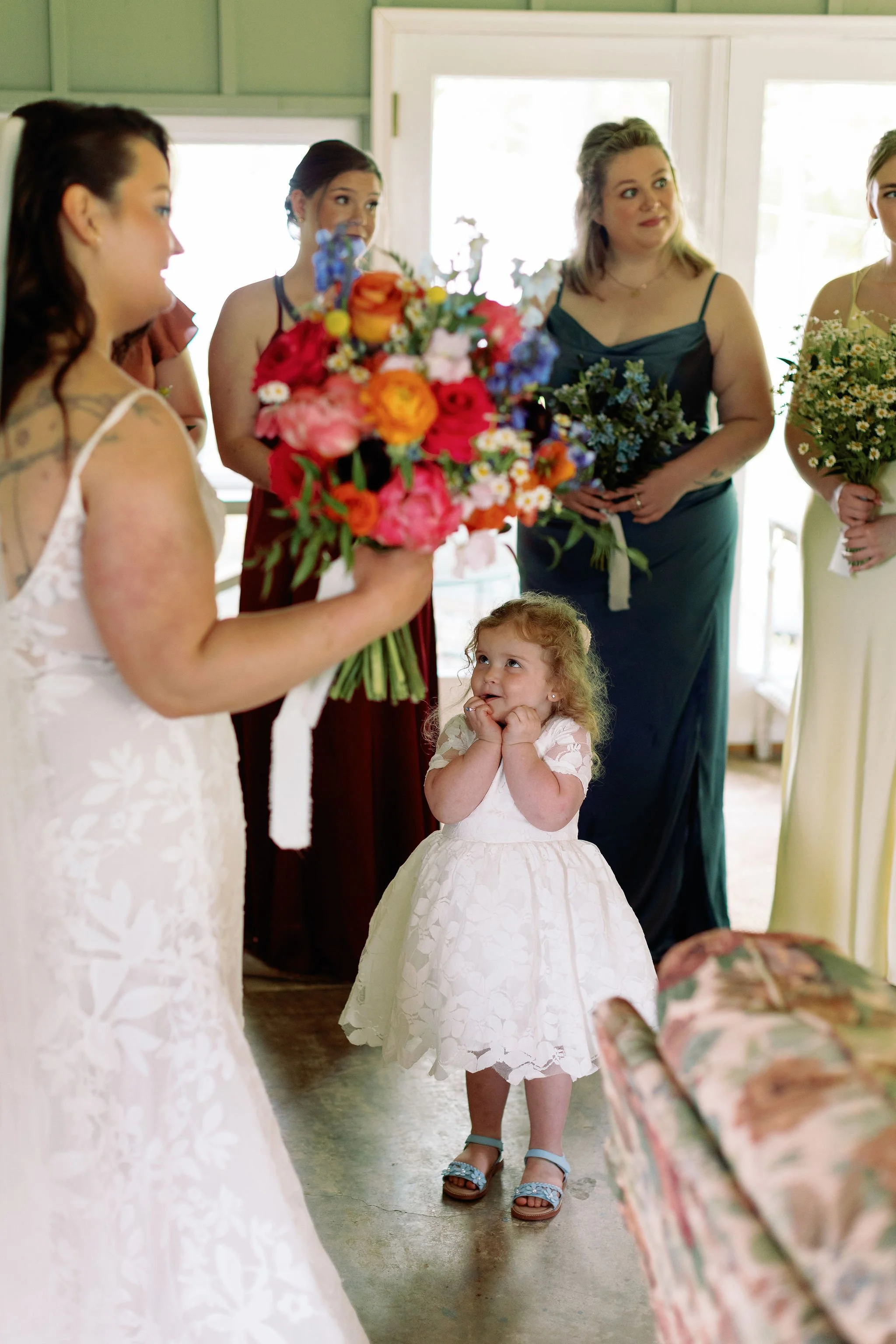A young girl with curly hair wearing a white dress and sandals standing with hands on her cheeks, looking at a woman holding a large bouquet of flowers during a wedding celebration.